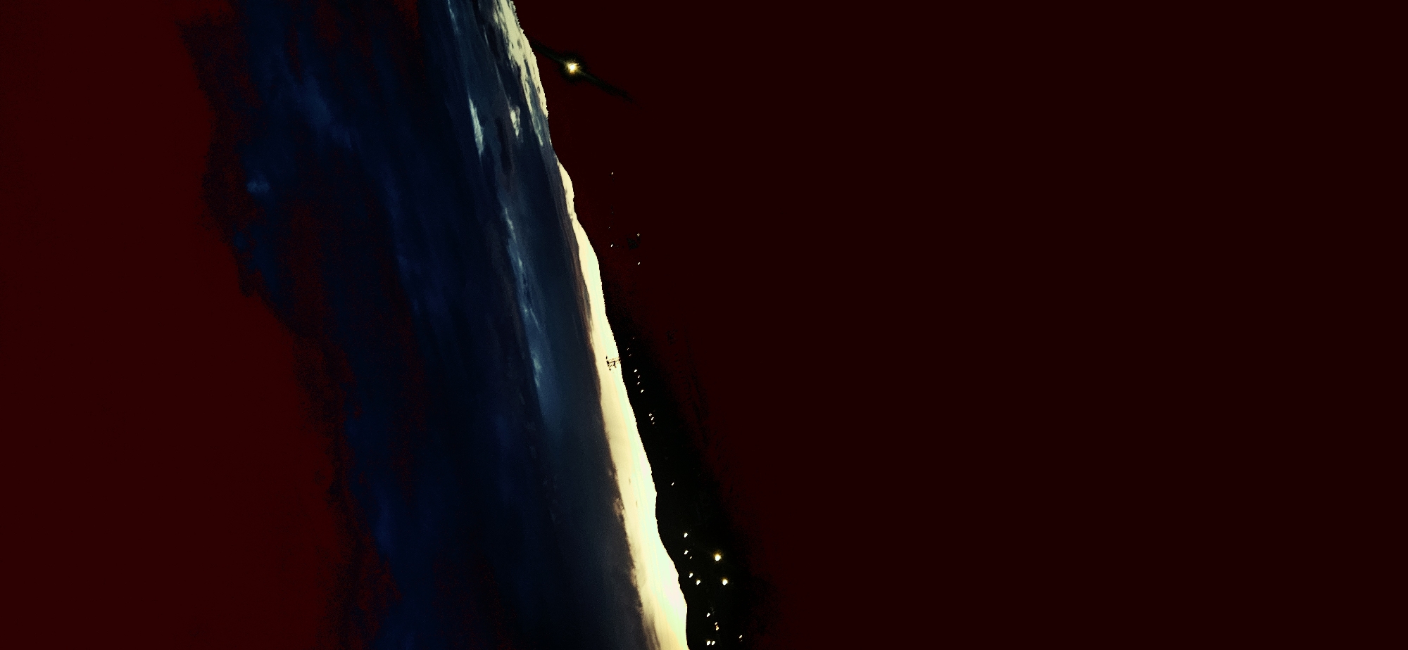 The image presents a dark, high-altitude or orbital view, likely taken at night or during deep twilight, looking down at a terrestrial landscape. A dominant vertical band occupies the left and central parts of the frame, showing varied textures of dark blue and lighter shades, culminating in a bright, almost white and yellow-gold illuminated edge on its right side. This band appears to be either a significant cloud formation or a mountain range.
To the right of this bright edge, the scene transitions into a deep reddish-brown expanse, resembling a dark sky or shadowed terrain. Scattered throughout this dark area, particularly along the lower portion of the bright band, are numerous small, distinct yellow-orange lights, indicating a city or settlement at night.
A notable object in the upper central area is a dark, elongated form with a bright yellow light at its front, characteristic of an aircraft or spacecraft in flight. Further down the bright illuminated edge, a smaller, illuminated, somewhat geometric structure is visible, possibly a building or tower.
The overall impression is one of vastness and mystery, contrasting natural geological or atmospheric features with human activity represented by the artificial lights and the flying object. Given the context, this could be an aerial view of the region around Momin Prohod, Bulgaria, at night. No visible text is present.