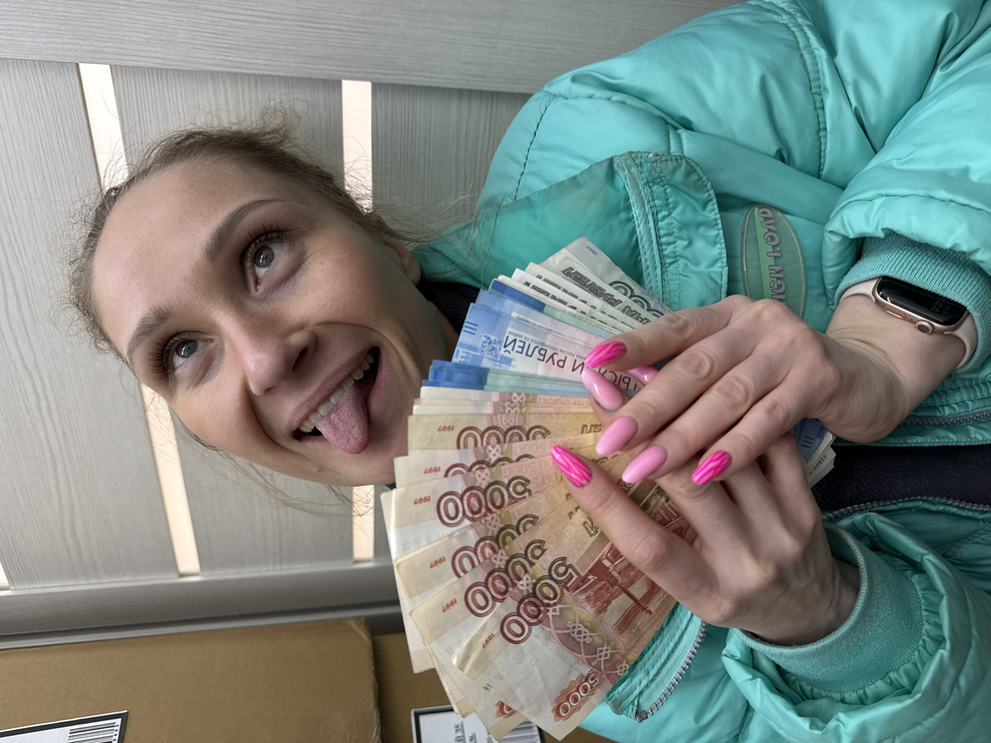 An indoor, eye-level portrait shows an adult female in Chaikovsky, Russia, centered, holding a fanned stack of Russian ruble banknotes. She exhibits a broad smile, extended tongue, and is looking upwards and to her left. Her light-brown hair is pulled back, and she wears a turquoise puffer jacket over a dark garment, with a light-colored smartwatch on her left wrist. Her fingernails are manicured with a combination of light pink and vibrant fuchsia polish, some featuring a striped design. The banknotes are predominantly 5000-ruble denominations (red/brown) and several 2000-ruble denominations (blue/green). The background consists of light brown cardboard boxes stacked on the left, one displaying a white label and barcode, and a light-colored paneled door or wall to the right.