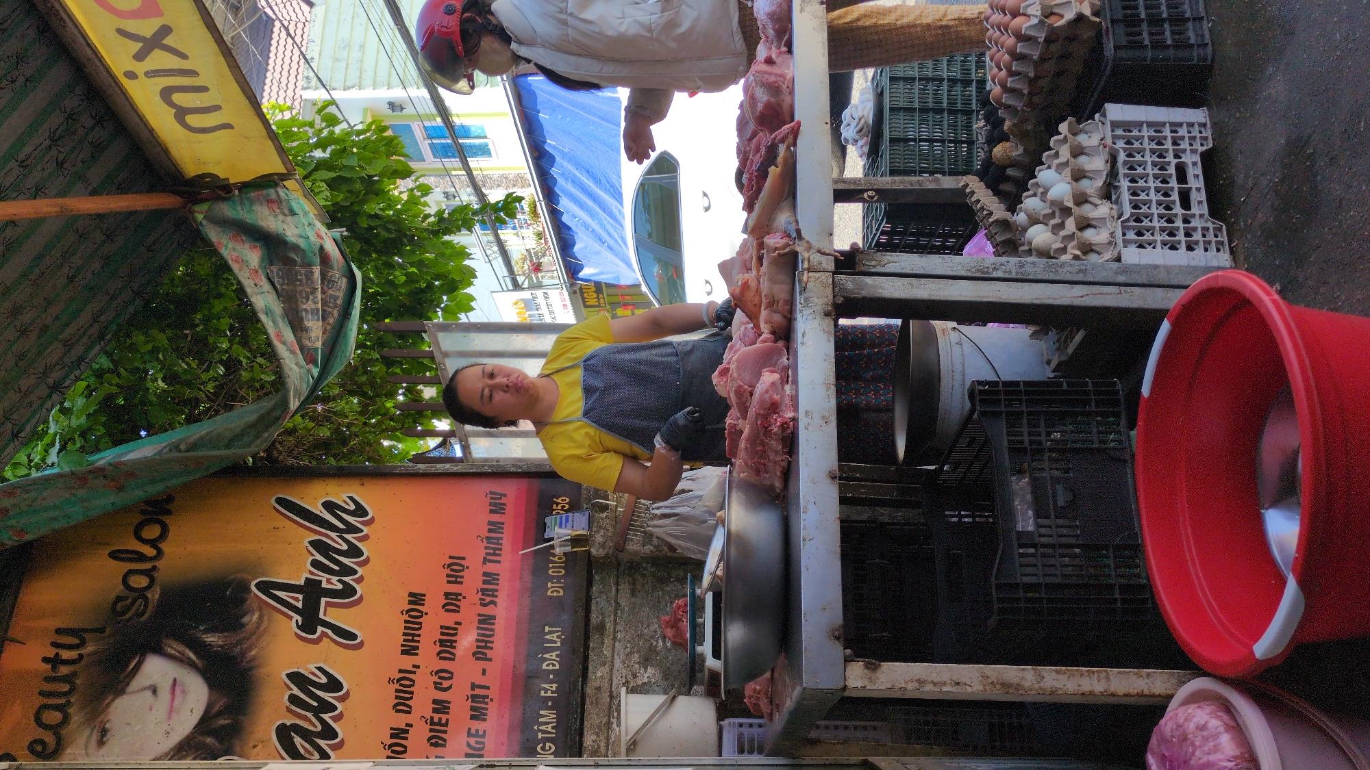 The image shows a street vendor in Da Lat, Vietnam, selling fresh pork. The vendor, wearing a yellow shirt and grey apron, stands behind a metal table covered in raw meat. Behind the vendor, on a shelf, are crates of eggs and other containers. Another person, wearing a white jacket and helmet, is also visible near the meat. To the left is a sign advertising a beauty salon named "An Anh," which offers services like hair styling, makeup, and tattooing. The sign indicates the salon is located in the F4 area of Da Lat. A yellow sign with "mix" is also on the left.