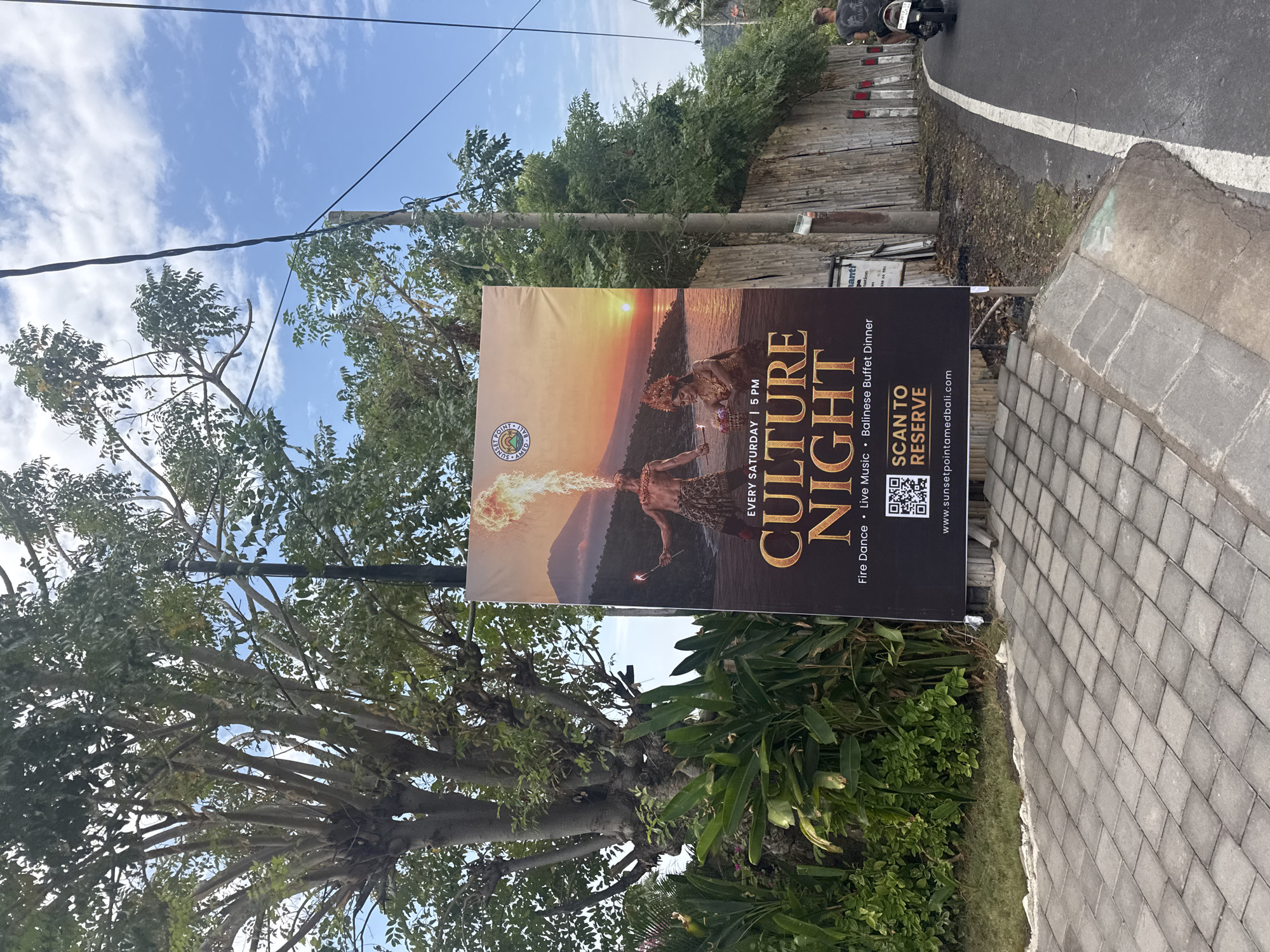 An outdoor advertisement banner for "Culture Night" is displayed on a pole alongside a paved walkway and a road. The banner features an image of two performers at sunset, one breathing fire and another in traditional attire. Text on the banner reads "EVERY SATURDAY | 5 PM", "CULTURE NIGHT", "Fire Dance • Live Music • Balinese Buffet Dinner", and "SCAN TO RESERVE www.sunsetpointamedbali.com". The surrounding environment includes lush green foliage, trees with exposed branches, a bamboo fence, and a clear blue sky with scattered clouds. In the background, a person is riding a motorcycle on the road. The scene is set in Abang, Indonesia, as indicated by the website address.