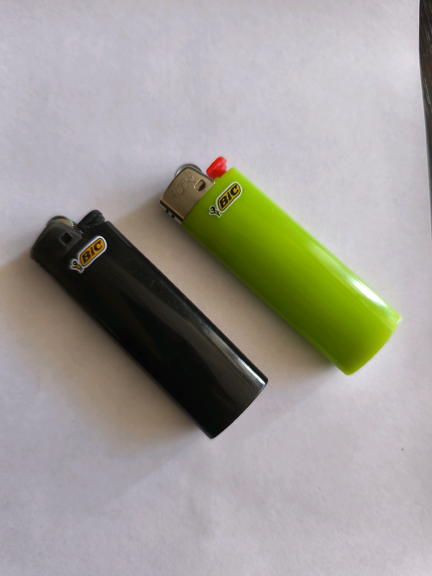 Two BIC lighters are placed on a white, textured surface. One lighter is black, and the other is bright green. Both lighters have the BIC logo with a small bee graphic on them. The black lighter is positioned to the left and slightly below the green lighter. The green lighter is oriented at an angle, with its red ignition mechanism visible at the top. The surface appears to be a piece of paper or fabric, with subtle shadows cast by the lighters. The lighting is even, with no strong directional sources, suggesting an indoor setting or diffused daylight. There is no indication of the specific location within Karachayevsk, Russia, as the image is a close-up of the objects and their immediate surroundings.