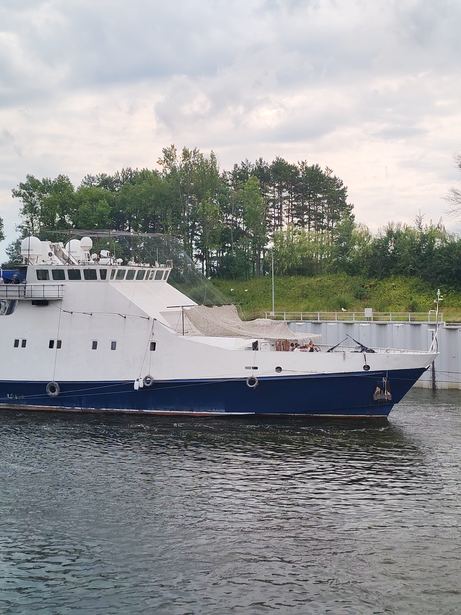 A white and blue vessel is docked in the waters of Bol'shie Mkhi, Russia. The ship has a white superstructure with multiple windows, radar dishes, and antennas. A blue hull is visible below the waterline. The vessel is positioned alongside a concrete structure, possibly a dock or canal wall. Trees and foliage line the background, with a cloudy sky overhead. Several people are visible on the deck of the vessel.