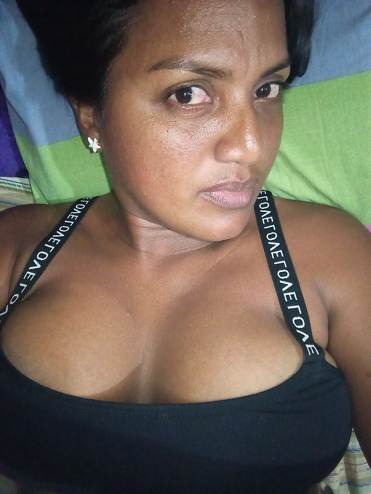 A close-up, eye-level photograph captures a person, likely a woman, with a dark complexion and dark hair, reclining indoors. The subject is looking directly at the camera with a neutral expression. Her dark hair is pulled back, revealing a small, white, flower-shaped earring on her left ear. Her dark eyes are prominent, with the left eye showing noticeable redness. She has full lips and defined eyebrows. Her skin appears somewhat dewy or oily.
She is wearing a fitted black top or bra with wide shoulder straps. The straps feature a repeating pattern of white, capitalized letters against the black fabric. The visible text reads "ΣΤΟΛΕΓΤΟΛΕΓΤΟΛΕΓΤΟΛΕΓΤΟΛΕΓΤΟ", where "ΣΤΟΛΕΓ" (Greek letters for 'STOLEG') is a repeating pattern of white capitalized characters. The top reveals cleavage.
The background consists of soft, patterned fabric, likely pillows or bedding, displaying horizontal stripes in shades of green, blue, and purple. The lighting is diffused, highlighting the subject's features. The scene suggests a private, relaxed setting, possibly a bedroom.