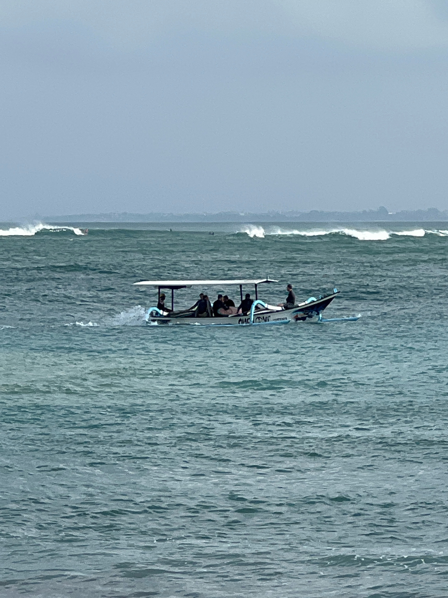 A traditional Balinese outrigger boat, painted white and blue, carries five individuals across choppy, teal-colored ocean waters under an overcast sky. The boat has a simple canopy providing shade. The individuals appear to be seated and observing the water or engaged in conversation. In the background, the ocean features significant waves breaking, suggesting a surf spot. A hazy, indistinct shoreline is visible in the far distance, likely representing the coast of Kuta, Indonesia. The overall scene depicts a cloudy day at sea near a popular surfing destination.