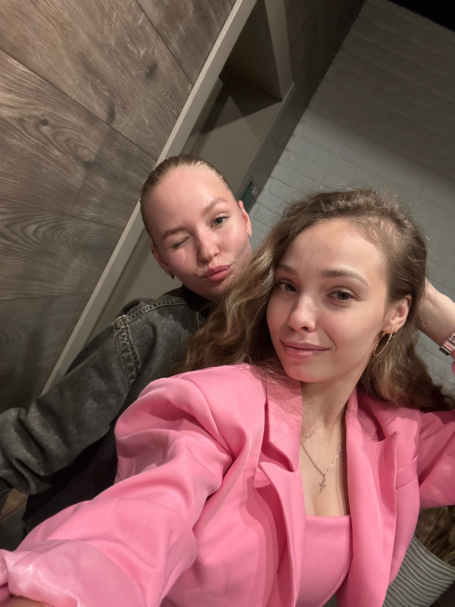 The image shows two young women, appearing to be in their late teens or early twenties, posing for a selfie indoors. The woman in the foreground, on the right, has long, wavy brown hair and is wearing a bright pink blazer over a matching top and a cross necklace. She has a gold hoop earring in her visible ear. The woman behind her, on the left, has her blonde hair pulled back and is wearing a dark grey jacket. She is winking and puckering her lips. The background appears to be a modern interior with wooden paneling on the left and a brick-patterned wall on the right. There is no identifiable information within the image to suggest it is located in Chaikovsky, Russia.