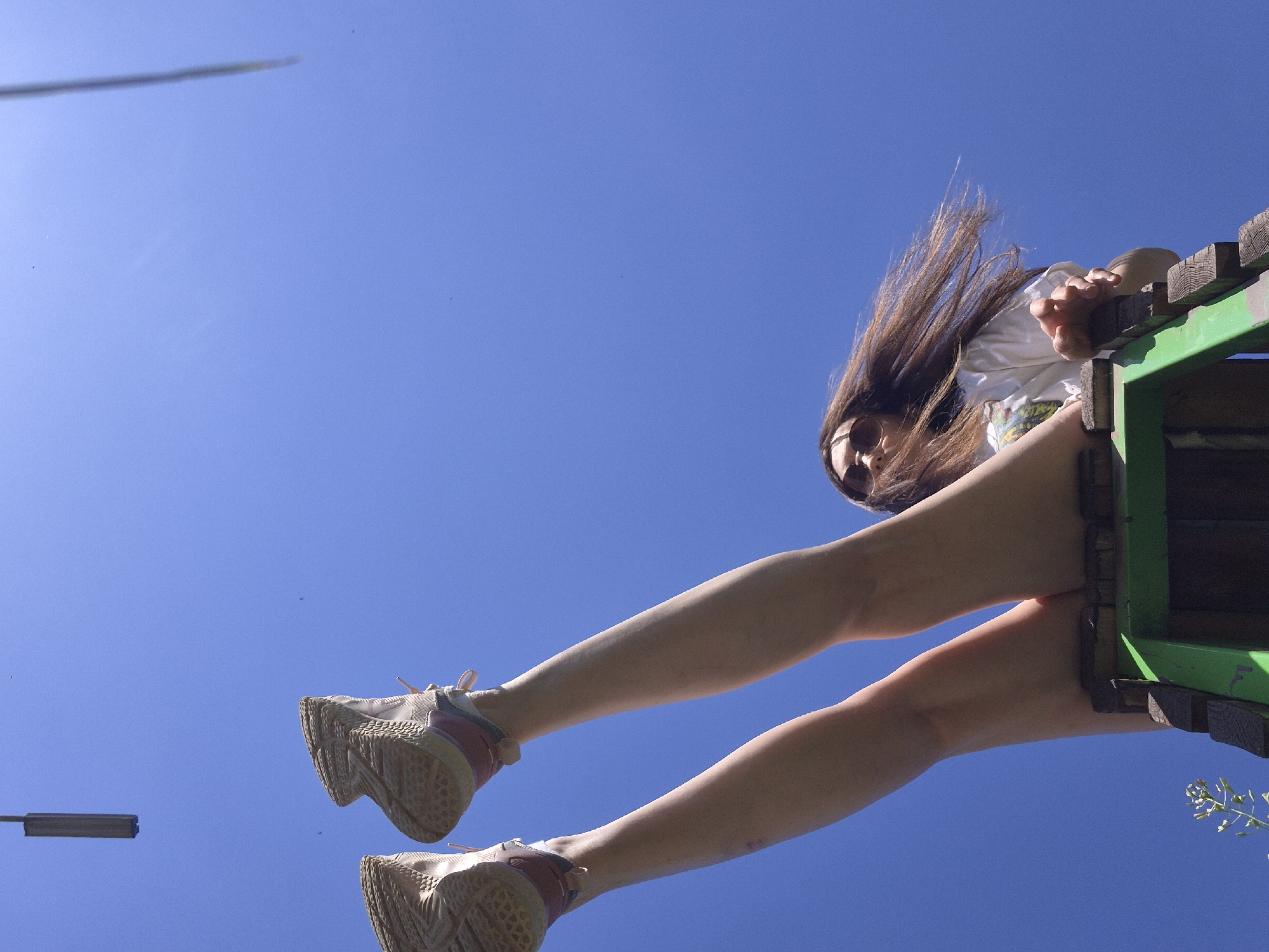 A low-angle shot shows a person hanging from a wooden structure, possibly a treehouse or playground equipment. The subject is wearing a white shirt and athletic shoes, with long brown hair partially obscuring their face. The background is a clear blue sky, suggesting a sunny day. The wooden structure is painted green and appears to be in an outdoor setting. A light pole can be seen in the bottom left hand corner of the image. Location: Unknown, Unknown.