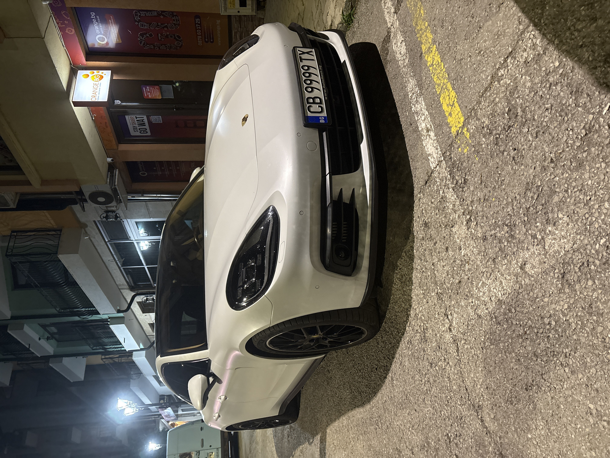 A white Porsche Panamera is parked on an asphalt street at night. The car is facing slightly towards the right of the frame. It has a black roof and black wheels. The license plate is visible and reads "CB 9999 TX". In the background, there is a building with shops. One shop has a sign that says "ORANGE.BG" and another has a sign that says "STAY SHARP GO WAT". There is also a street lamp visible on the left side of the image. The ground is marked with yellow and white lines.