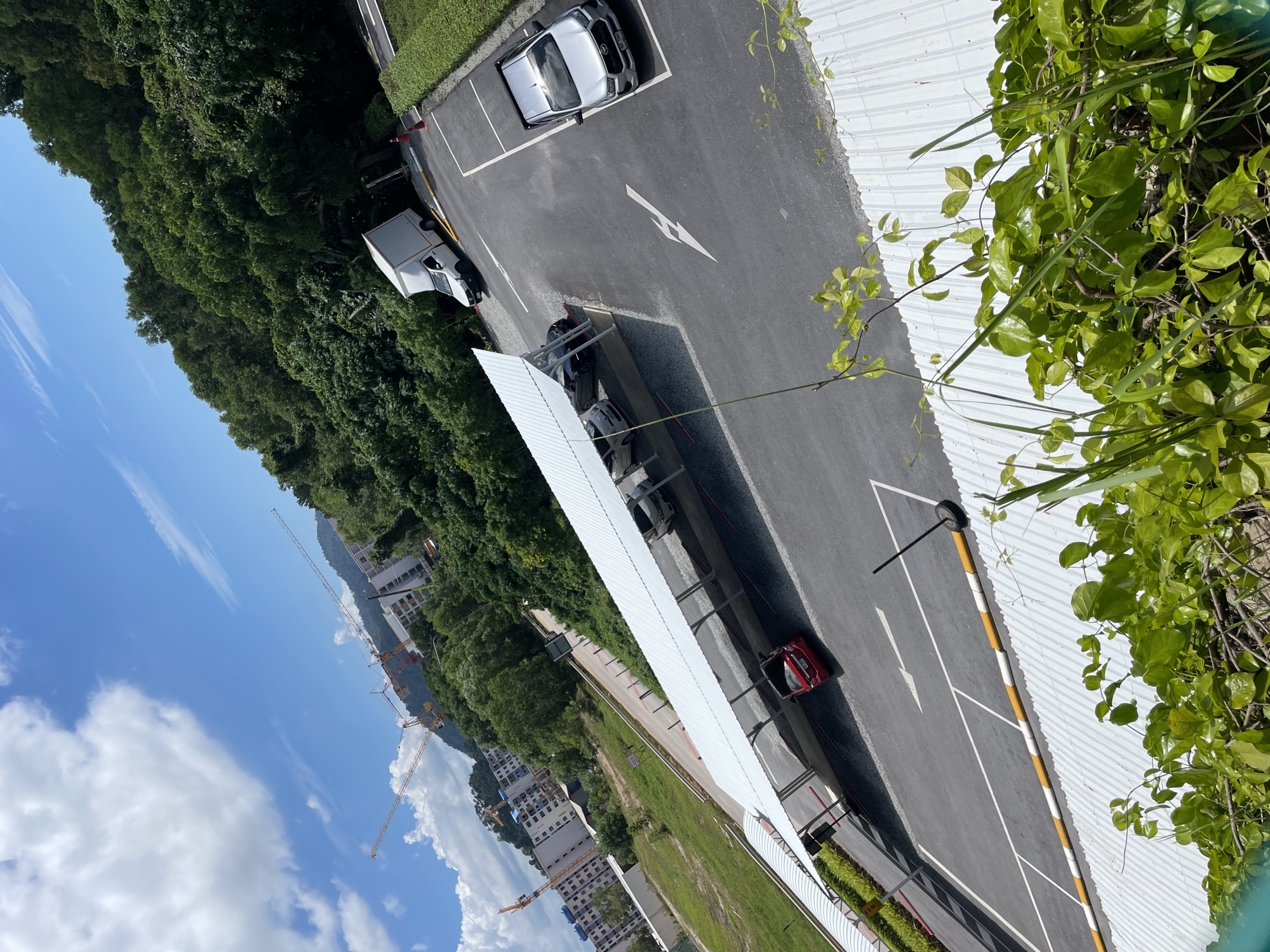 The image depicts an outdoor scene on a bright, sunny day, likely in a developing area. In the foreground, lush green foliage and vines partially obscure a corrugated metal roof. Beyond this, a paved parking area is visible, featuring marked parking spaces and directional arrows. Several vehicles are parked in the lot, including a silver SUV, a white box truck, and a red car parked under a covered structure. This covered structure, with a white, translucent roof supported by metal beams, appears to be a carport or an open-air parking shelter.
In the mid-ground, the landscape transitions to a green, grassy area bordering a road. Further in the background, a densely wooded hillside rises, covered in vibrant green trees. In the distance, buildings under construction are visible, with several tall construction cranes actively working against a bright blue sky dotted with white clouds. The overall impression is one of a partially developed urban or suburban environment undergoing construction, set against a backdrop of natural greenery.