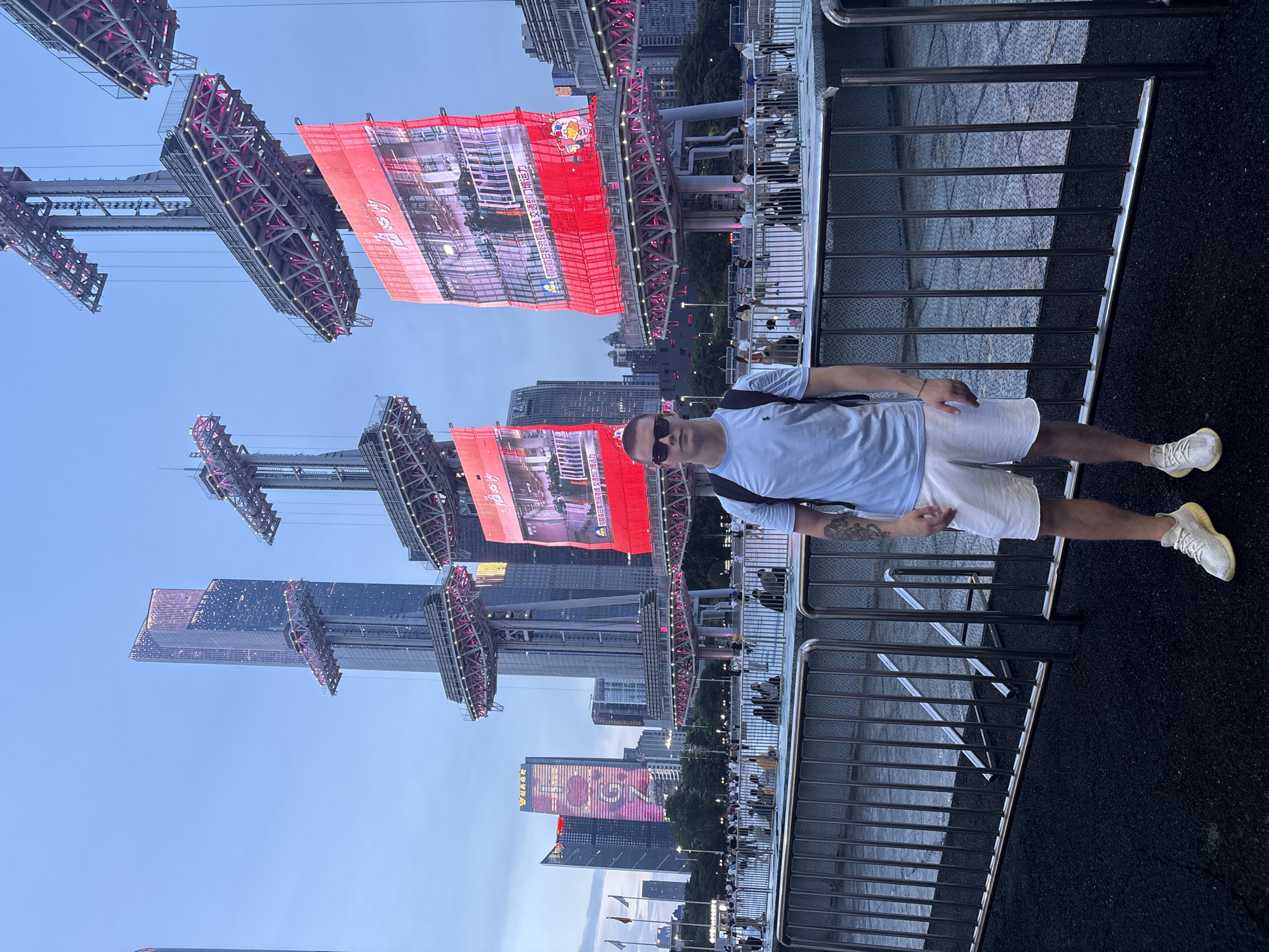 This is a vertical, eye-level photograph taken outdoors in an urban setting. The main subject is a young man wearing a light blue t-shirt, white shorts, sunglasses, and white sneakers. He has tattoos on his left arm and a red string bracelet on his left wrist. He is standing on a dark asphalt surface next to a metal railing.
In the background, a cityscape with tall buildings is visible. Several large, illuminated structures with red banners hang from them, displaying images and Chinese text. These appear to be part of an outdoor event or advertisement. The sky is clear and blue, suggesting it is daytime, likely late afternoon or early evening. The lighting on the buildings and banners indicates artificial illumination.
The scene appears to be a public area, possibly a pedestrian walkway or viewing platform, overlooking the city. The presence of a railing and a crowd of people visible in the distance suggests this is a popular spot. The overall mood is casual and relaxed, with the man looking towards the camera.
Visible text includes Chinese characters on the red banners. One banner prominently displays a Hello Kitty character. Another banner shows large pink numerals that appear to read "100". The image captures a vibrant urban environment with a focus on modern architecture and commercial displays.