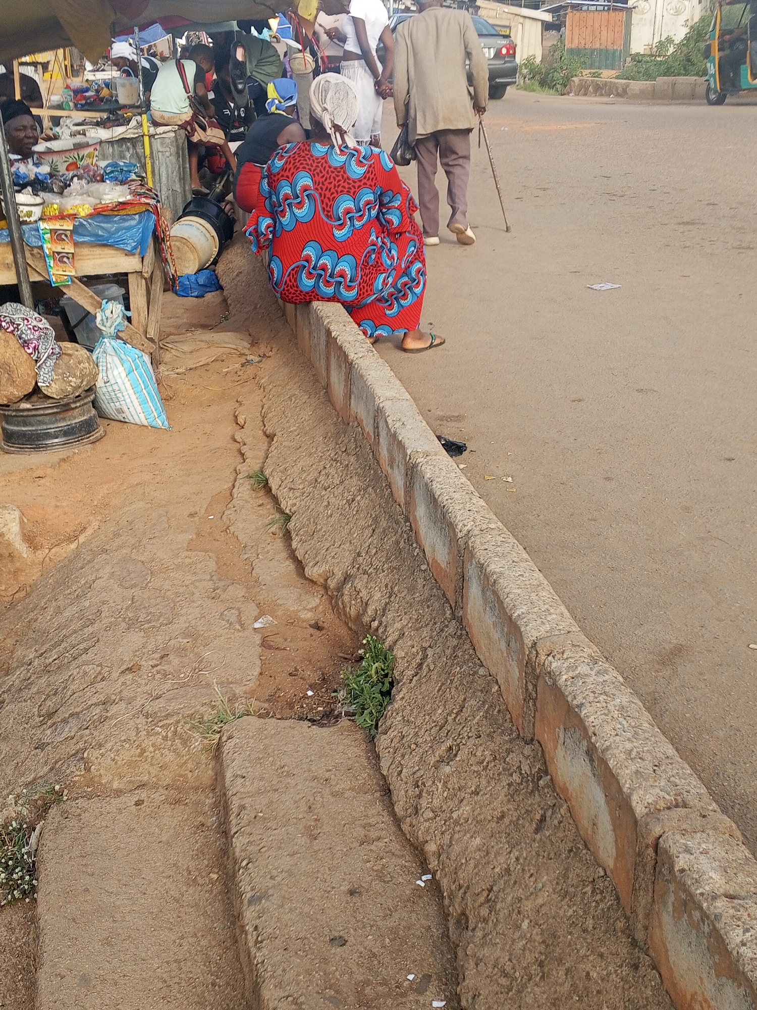 The image depicts a roadside scene in Gwong, Nigeria. In the foreground, a woman in a bright red and blue patterned dress is sitting on a low concrete barrier that separates the road from a lower, eroded dirt area. The barrier runs parallel to the road and has patches of green vegetation growing at its base.
Behind the woman, on the left side of the frame, is a makeshift market stall beneath a tarpaulin, with a vendor sitting among various goods for sale. Assorted items and containers are visible on the stall, including bags and drums.
On the road, an elderly man with a walking stick is walking away from the camera. A car is partially visible behind him. Other individuals can be seen near the market stall, engaged in various activities. The environment appears to be a bustling commercial area.