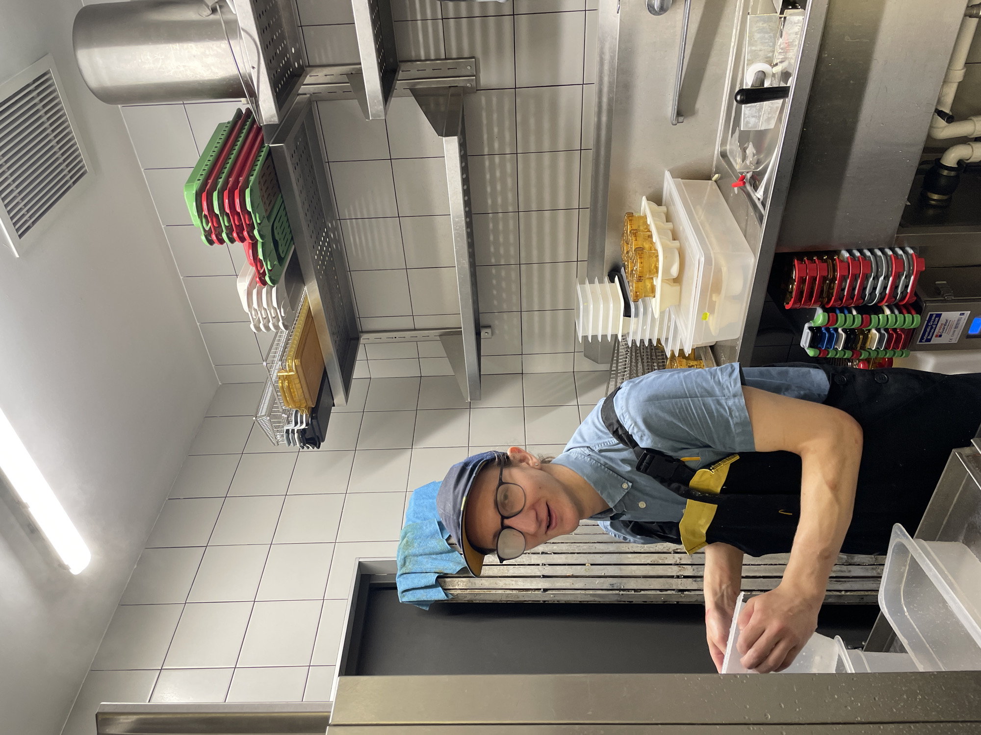 A person wearing a blue button-down shirt, black overalls with a yellow bib, a baseball cap, and glasses stands in a kitchen and holds up a clear plastic container. The person is looking at the camera and smiling. The kitchen has tiled walls and stainless steel shelves that hold stacked red and green plastic containers, along with a large metal pot. There are also stacks of white containers and a tray of small yellow containers on a counter. A sink is visible to the right, and a black and silver appliance is on the floor.