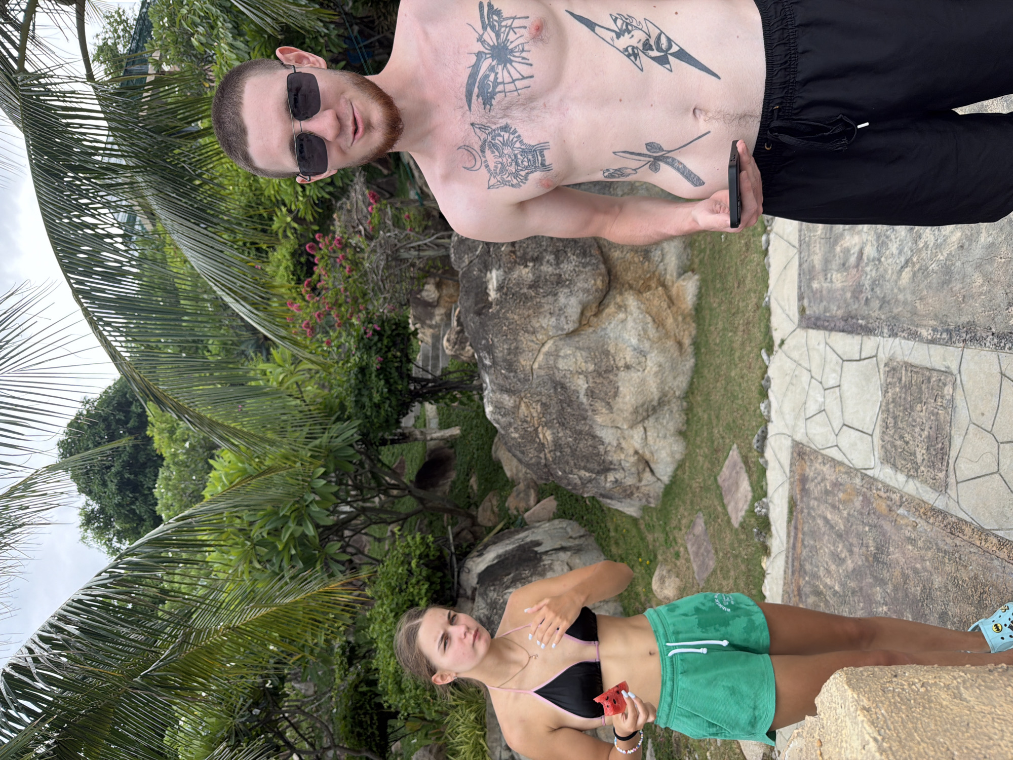 Two individuals, a young woman and a young man, are standing outdoors in what appears to be a tropical or semi-tropical environment. The woman, on the left, is wearing a black bikini top with pink trim and bright green shorts. She is holding a slice of watermelon in her right hand and has her left hand near her chest. She has a necklace on and several bracelets on her left wrist. The man, on the right, is shirtless and wearing black swim trunks. He has multiple tattoos on his chest and abdomen, and is wearing dark sunglasses. He is holding a smartphone in his right hand.
The background consists of lush green vegetation, large rocks, and palm trees, suggesting a location with a warm climate. The ground beneath them is paved with stone tiles. The presence of palm trees and abundant greenery suggests this location could be within the city of Bắc Nha Trang, Vietnam, known for its beaches and tropical landscapes. The overall scene depicts a casual, outdoor setting, possibly a resort or park.