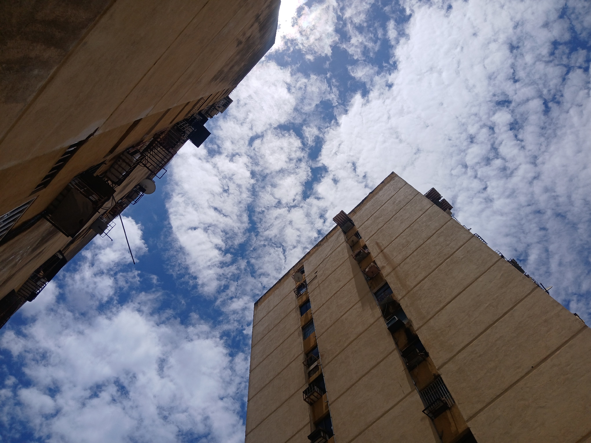 A low-angle shot of two tall, rectangular beige buildings under a sky with fluffy white clouds. The buildings, likely apartment complexes, appear to be of a similar design, featuring vertical lines and small balconies. The angle suggests the photo was taken from a narrow street or alley between the buildings. The location is Ciudad Guayana, Venezuela.