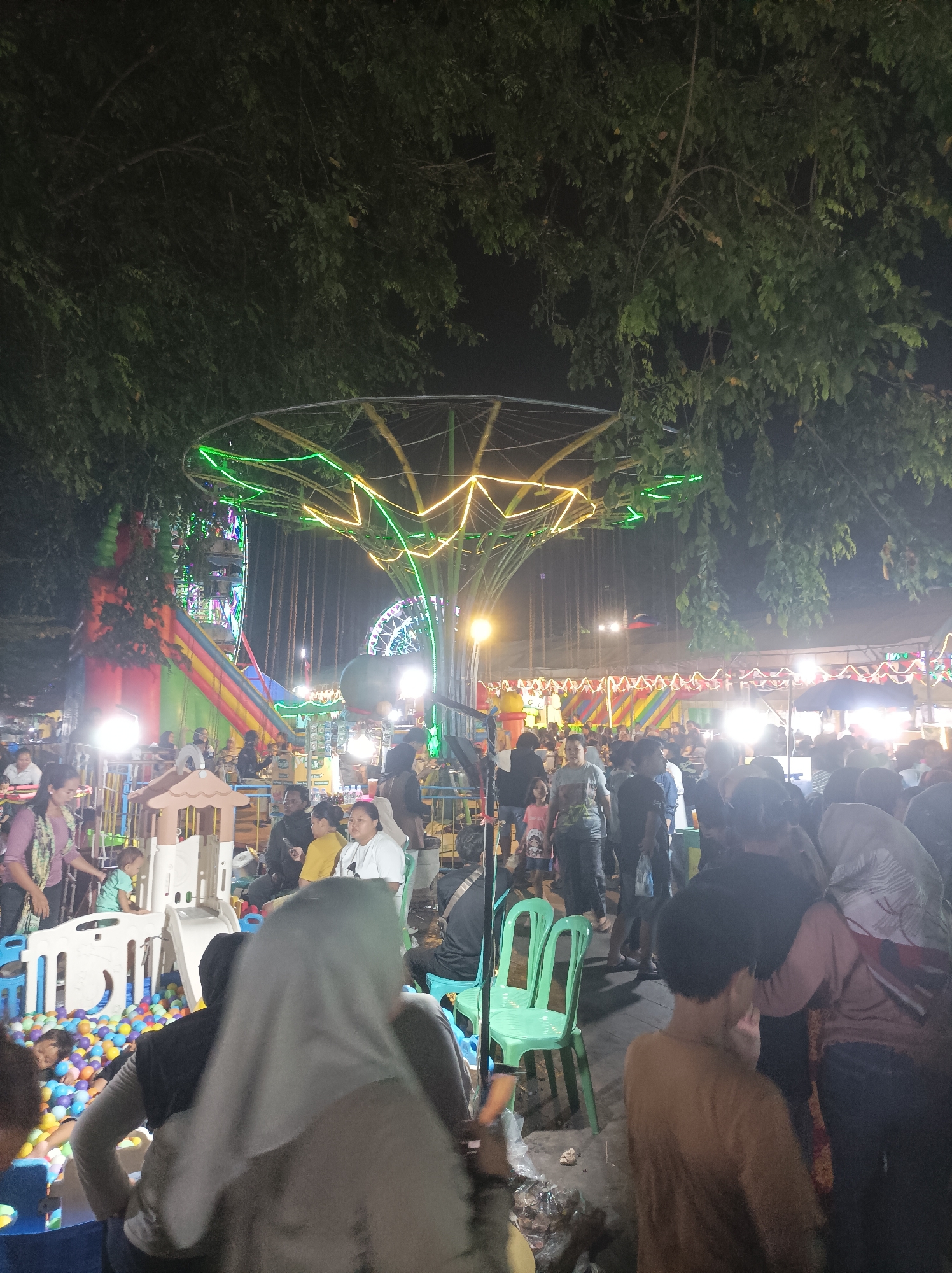 The image depicts a night scene at a bustling carnival or fairground, likely in the city of Cirebon, Indonesia, given the context. A large, illuminated ride, possibly a swing ride or carousel, dominates the upper portion of the frame, adorned with bright green and yellow lights. Below, a crowd of people of various ages are gathered, some standing, others seated. Several attractions are visible, including a large inflatable slide with colorful stripes, a ball pit with numerous colorful balls, and various stalls or booths. The environment appears to be outdoors, with trees providing a canopy overhead. The lighting is a mix of artificial carnival lights and general ambient illumination, creating a lively and somewhat chaotic atmosphere. People are dressed in casual attire, and the overall impression is one of a community event or local festival.