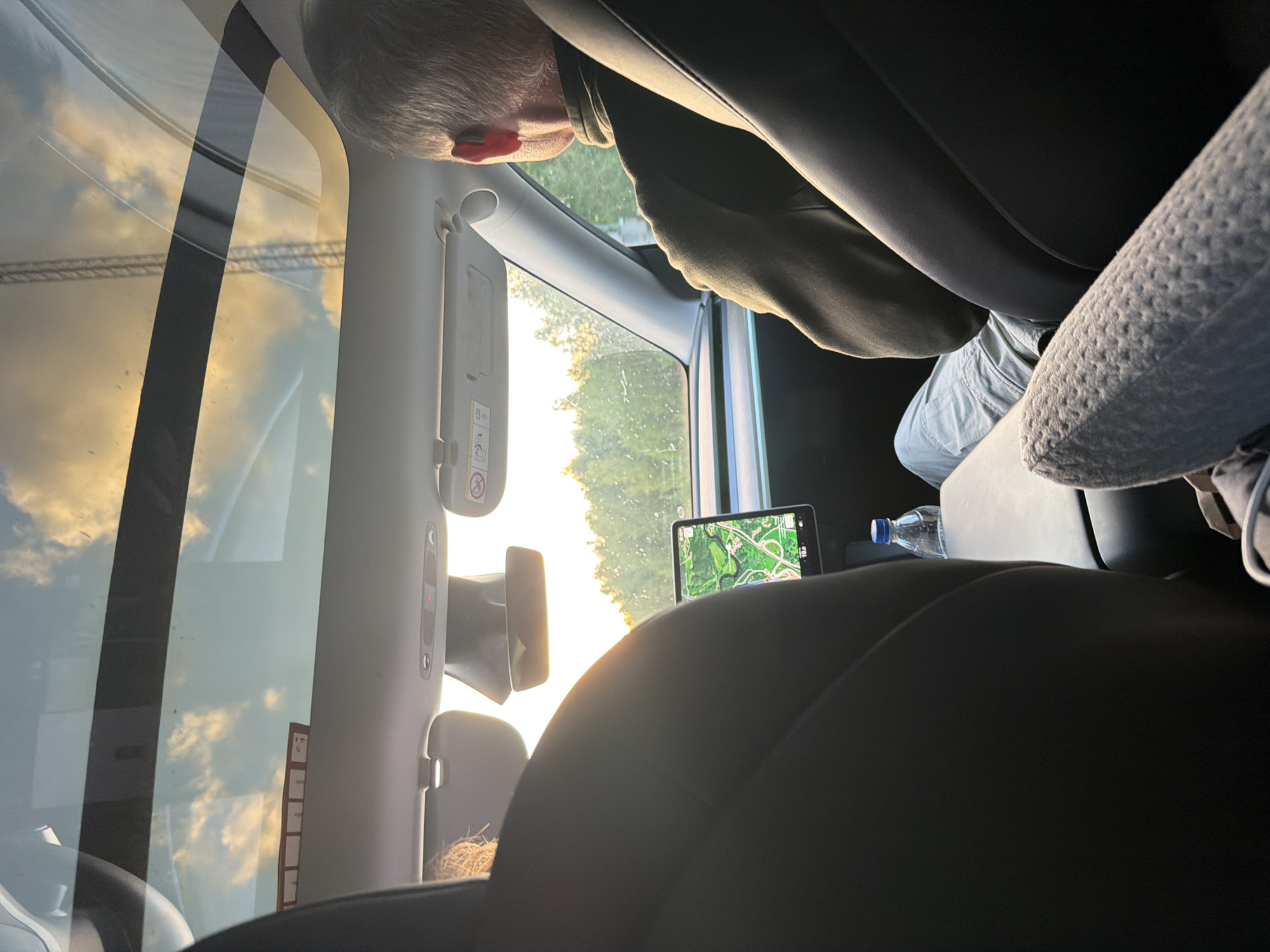 The image captures the interior of a car, likely a Tesla, with a panoramic glass roof showing a cloudy sky with hints of sunset colors. The view extends to the outside, revealing a treeline and what appears to be a body of water. In the foreground, two car seats are visible. In the driver's seat, a man with graying hair is seen from the side, wearing a dark jacket and light-colored pants. He has an earpiece in his right ear. In the passenger seat, a person's back is visible, with blonde hair peeking out. The car's touchscreen display is showing a map. A bottle of water sits on the center console. The image appears to be taken on a road, possibly on the E6 highway near Munkedal, Sweden, given the visible landscape and road signs indicating location.