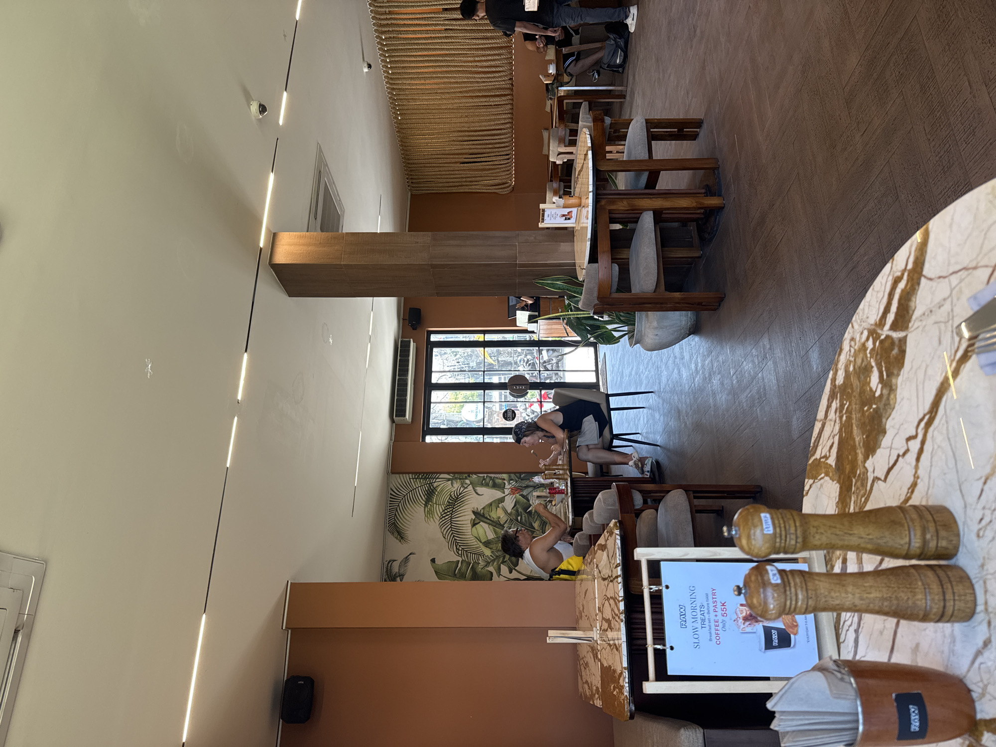 The image shows the interior of a restaurant in North Kuta, Indonesia. Two individuals are seated at a table, engaged in conversation while eating. The restaurant features brown walls, wooden accents, and a tropical-themed mural. Tables and chairs are arranged throughout the space, with a marble-topped table in the foreground displaying salt and pepper grinders, and a container of napkins. A menu board advertising "Slow Morning Treats" with coffee and pastry is also visible. In the background, a person stands near a doorway, and another person is seated at a table. The flooring is a dark wood-grain tile. Natural light enters from windows and is supplemented by overhead linear lighting.