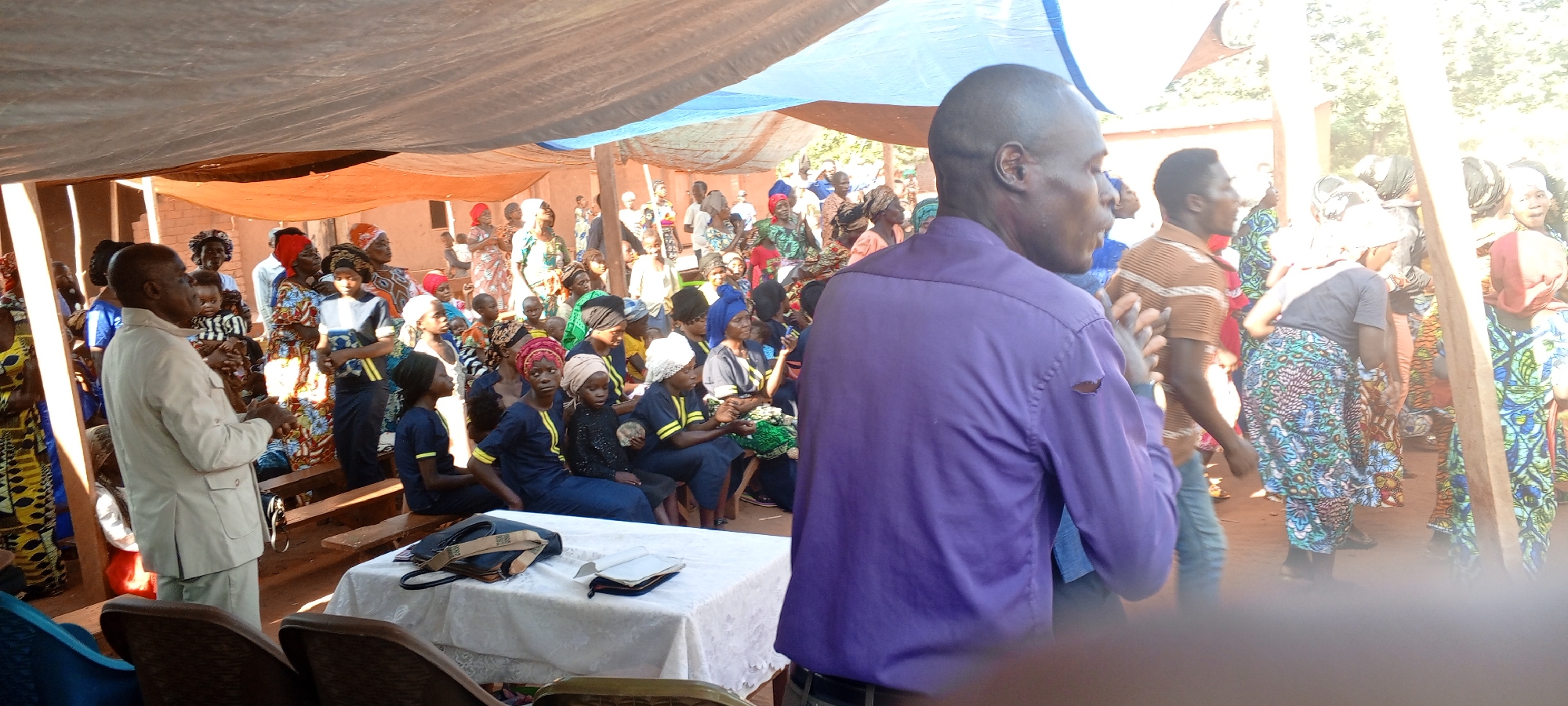 A large group of people is gathered outdoors under a makeshift canopy in Ngalula, Democratic Republic of the Congo. The crowd includes men, women, and children of various ages, many wearing colorful traditional clothing and headwraps. Some are seated on wooden benches, while others stand. A man in a purple shirt stands in the foreground with his back to the camera, clapping his hands. Another man in a light beige suit stands to his left. A table with a white tablecloth sits in front of the seated group, with a bag and a book on top. The environment appears to be a rural area with buildings visible in the background. The scene is brightly lit, suggesting daylight.
