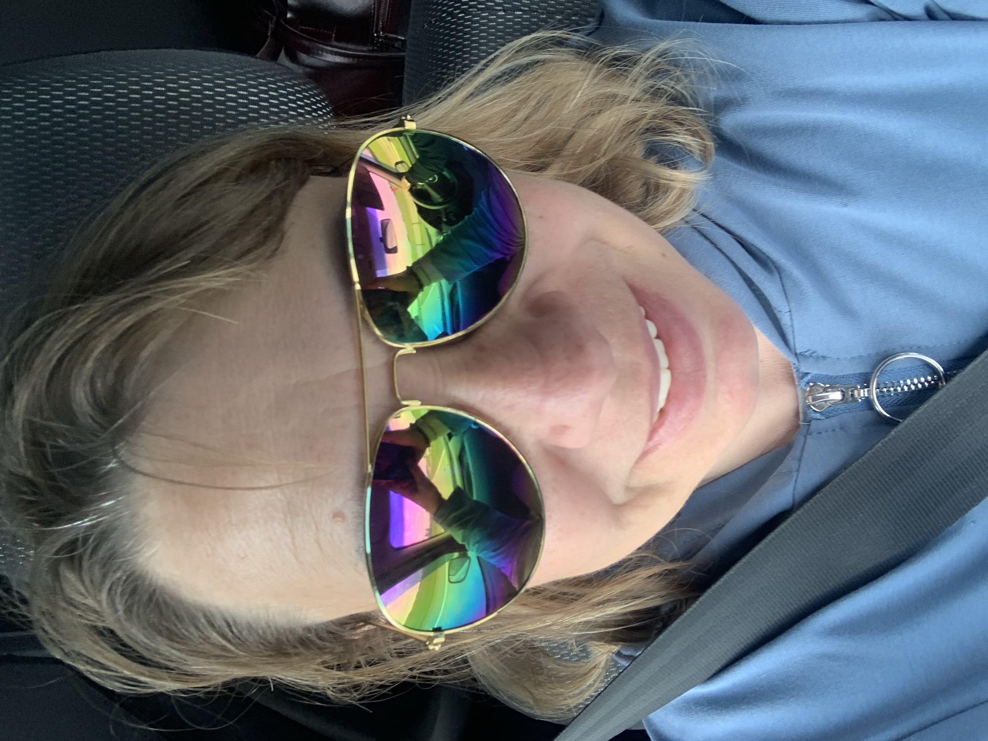 A woman with blonde hair and sunglasses is taking a selfie in a car. She is wearing a blue hooded jacket with a zipper. The sunglasses reflect the dashboard and the road ahead. A seatbelt is visible across her chest. The background shows a car seat. The image appears to be taken in the city of Azerski, Belarus.