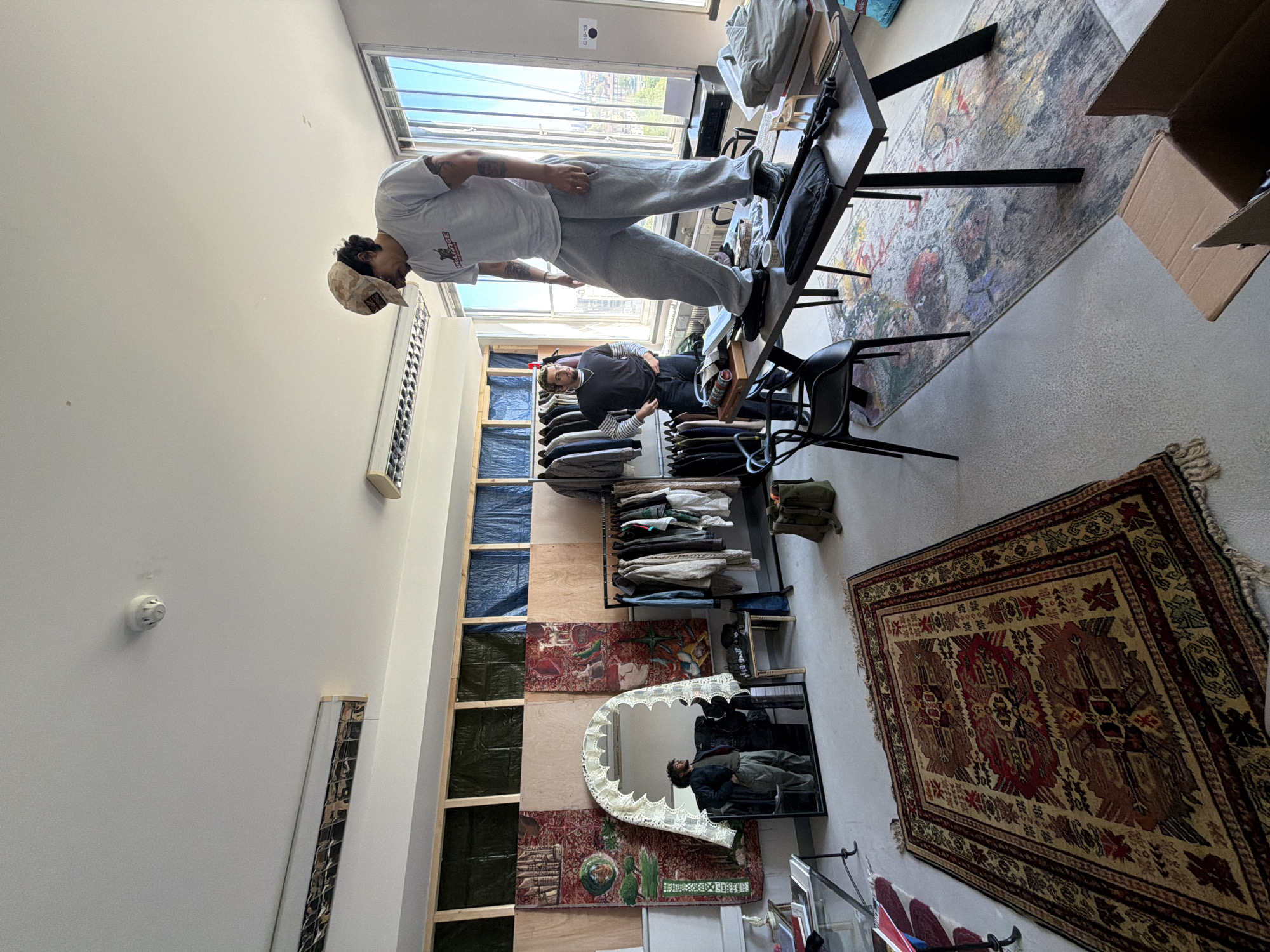 The image captures an indoor space, likely a studio or office, illuminated by bright daylight streaming through large windows that reveal blue sky and distant buildings. The scene is presented as if viewed from an overhead perspective due to the image's rotation, but people are oriented as if standing on the floor.
Two men are present. In the foreground, a man with tattooed arms, wearing a light-colored t-shirt with "BALENCIAGA" visible on it, grey sweatpants, and a camouflage baseball cap, stands casually on a desk. Behind him, a second man is seated at a different desk, wearing a dark long-sleeved shirt and dark trousers, looking towards the first man. His reflection is clearly visible in a large, ornate mirror with a white, lace-like frame.
The room is furnished with desks, an office chair, and shelving units displaying folded clothes and other items. The floor is covered by a large, colorful, abstract-patterned rug, while a smaller, traditional patterned rug hangs vertically on a wall. The walls also feature several unique tapestries or artworks. A cardboard box is visible near the floor rug. A fire alarm/smoke detector and vents are mounted on the ceiling. Text "C31-3" is visible on a wall near the windows. The overall atmosphere is casual and relaxed, suggesting a creative or informal workspace during the daytime.