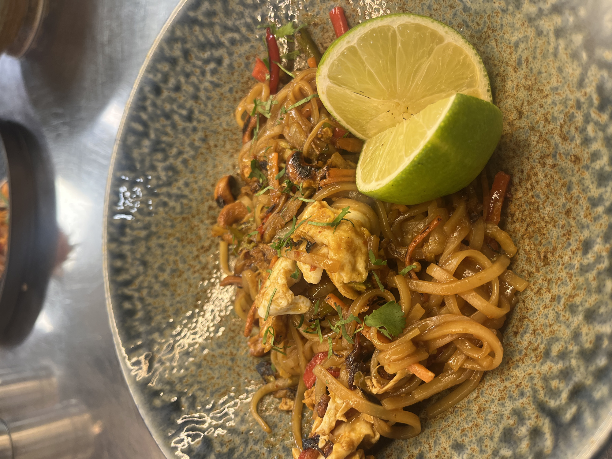 The image presents a close-up, overhead view of a prepared dish of stir-fried noodles, served in a distinctive ceramic bowl. The main subject is a hearty portion of wide, flat noodles, coated in a dark, savory sauce. Mixed into the noodles are various ingredients including pieces of cooked egg or omelet, julienned red bell peppers, and other vegetables that appear to be carrots and possibly onions. Several small, dark, oblong items resembling mussels or small clams are also visible, suggesting a seafood component to the dish.
The meal is garnished with freshly chopped green herbs, likely cilantro. Two bright green wedges of fresh lime are prominently placed on the rim of the bowl, ready to be squeezed over the food.
The serving bowl features a mottled, textured glaze, combining shades of blue-grey with rusty-brown speckles, giving it a rustic and artisanal appearance. To the left and top-left of the bowl, a reflective, metallic or stainless steel counter surface is visible, indicating a kitchen or restaurant setting.
No people are present in the image, and the primary activity depicted is the presentation of the prepared meal. The scene is well-lit, highlighting the colors and textures of the food. There are no discernible location cues within the image itself other than the provided context of Sofia, Bulgaria, and no visible text.