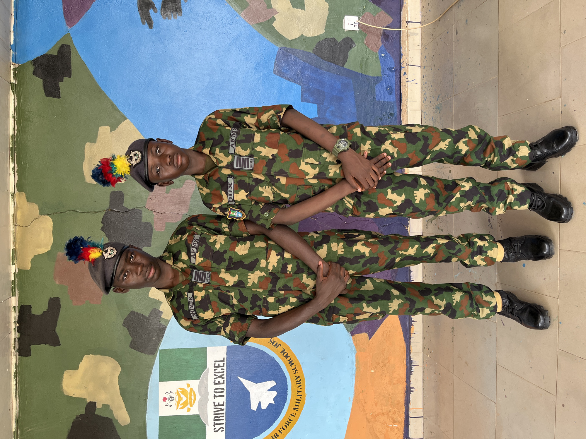 Two young males, dressed in military camouflage uniforms and dark berets with colorful plumes, stand facing the camera in front of a mural painted on a wall. The male on the left has a name tag that reads "SHIMFE" and another that reads "AFMS". The male on the right has a name tag that reads "DUX" and another that reads "AFMS". Both males have their hands clasped in front of them. The mural behind them is a camouflage pattern in shades of green, brown, and tan, with sections of blue and orange. A logo with the words "STRIVE TO EXCEL" and an image of a fighter jet is visible on the left side of the mural. The ground beneath them is tiled. The setting appears to be an outdoor area, possibly part of a military school or training facility. The image is taken at eye level. No specific location within the city of Bar Jirgi Summa, Nigeria is identifiable from the visual information present.