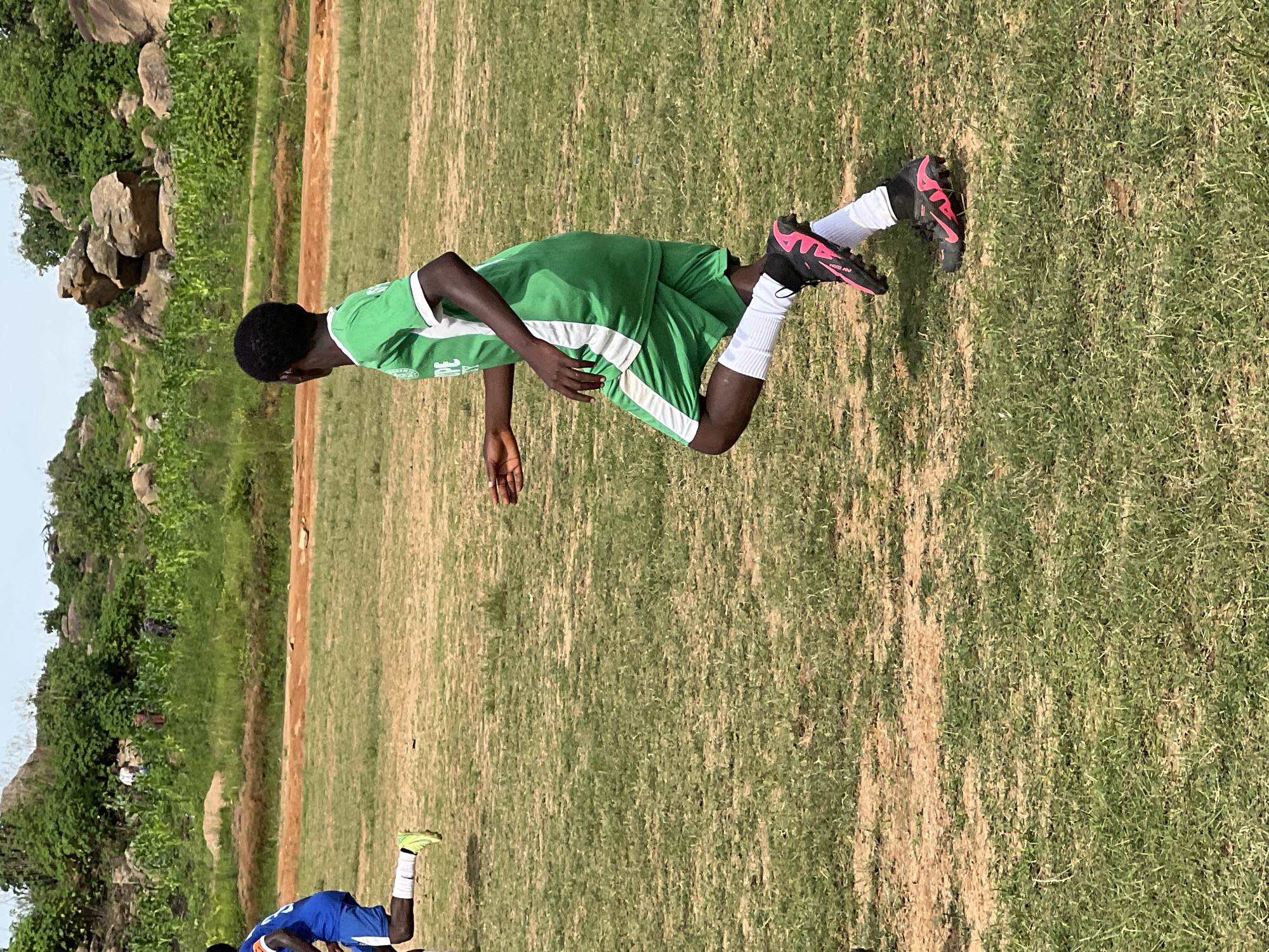 This image depicts a young person wearing a green soccer uniform with white trim, actively running on a grassy field. The soccer player's uniform consists of a short-sleeved jersey and shorts, paired with white socks and black soccer cleats accented with pink. The setting appears to be an outdoor sports field, likely in a rural or semi-urban area, given the backdrop of rocky hills, green vegetation, and what looks like a cultivated field of crops. There are no explicit markers within the image to pinpoint the exact location in Jos, Nigeria, beyond the general context of an open, natural environment suitable for soccer. The image captures a moment of athletic activity.