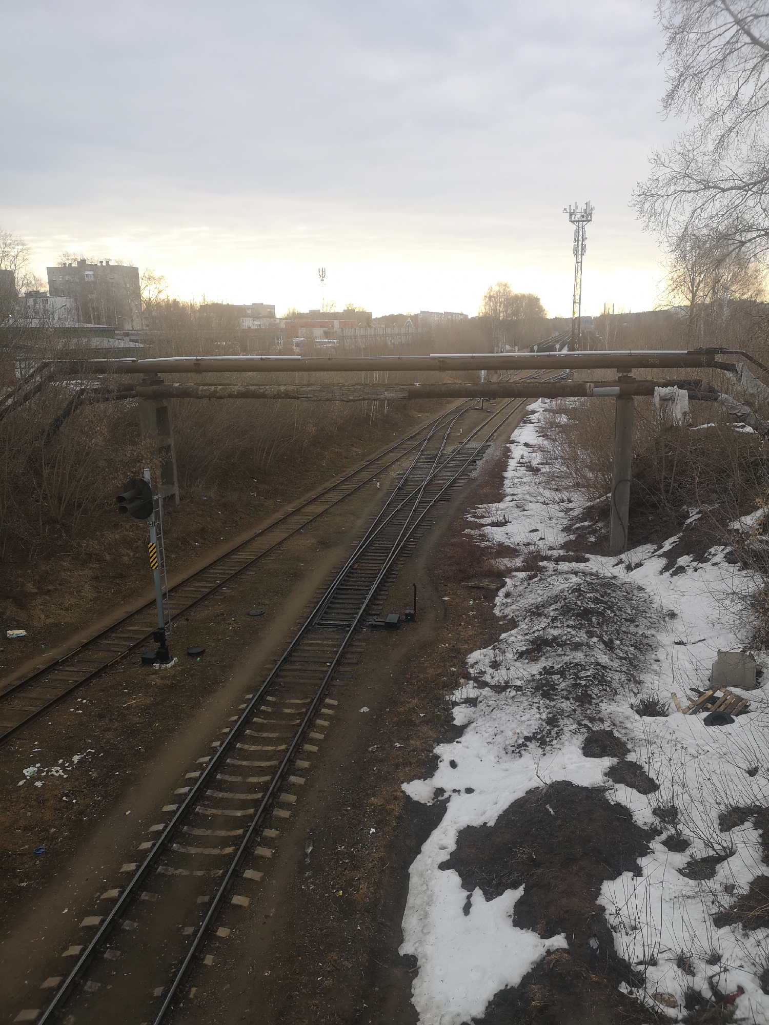 This image shows a railway scene in what appears to be a semi-industrial or urban fringe location. The focus is on a set of three parallel train tracks. The tracks are flanked by earth and vegetation. There is some remaining snow. A pipe structure, likely for industrial or utilities, spans across the tracks. A traffic signal for the rail line can be seen. Buildings and a cellular tower are visible in the background. Given the description, the location is the city of Chaikovsky, Russia. No people are visible in the image.