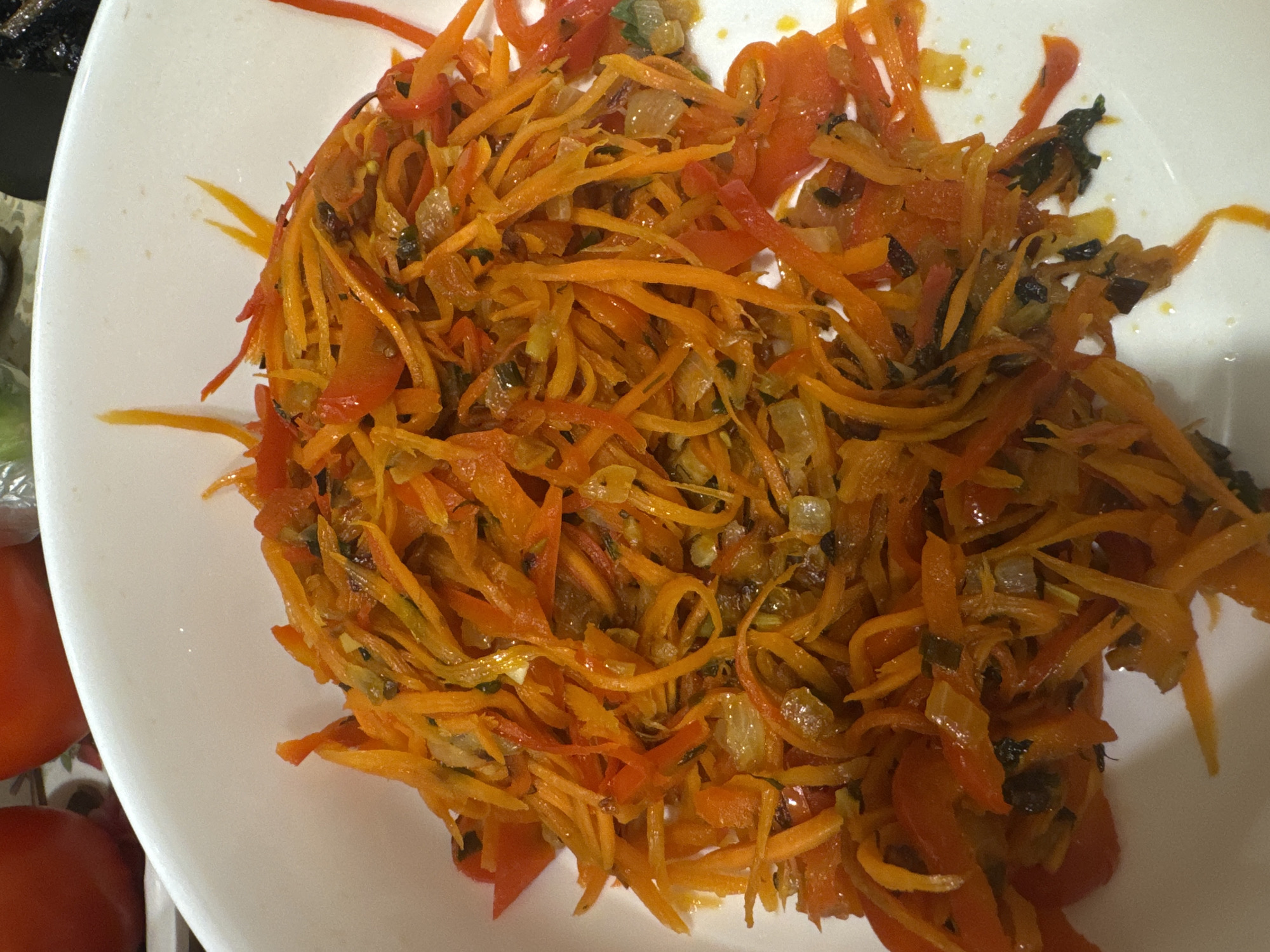 The image shows a plate of food, likely a salad, consisting primarily of julienned carrots and red bell peppers. There are also visible additions of what appears to be sautéed onions and some dark green herbs or spices. The food is presented on a white plate. The location is not discernible from the image itself. The image does not show any people, subjects, actions or interactions.