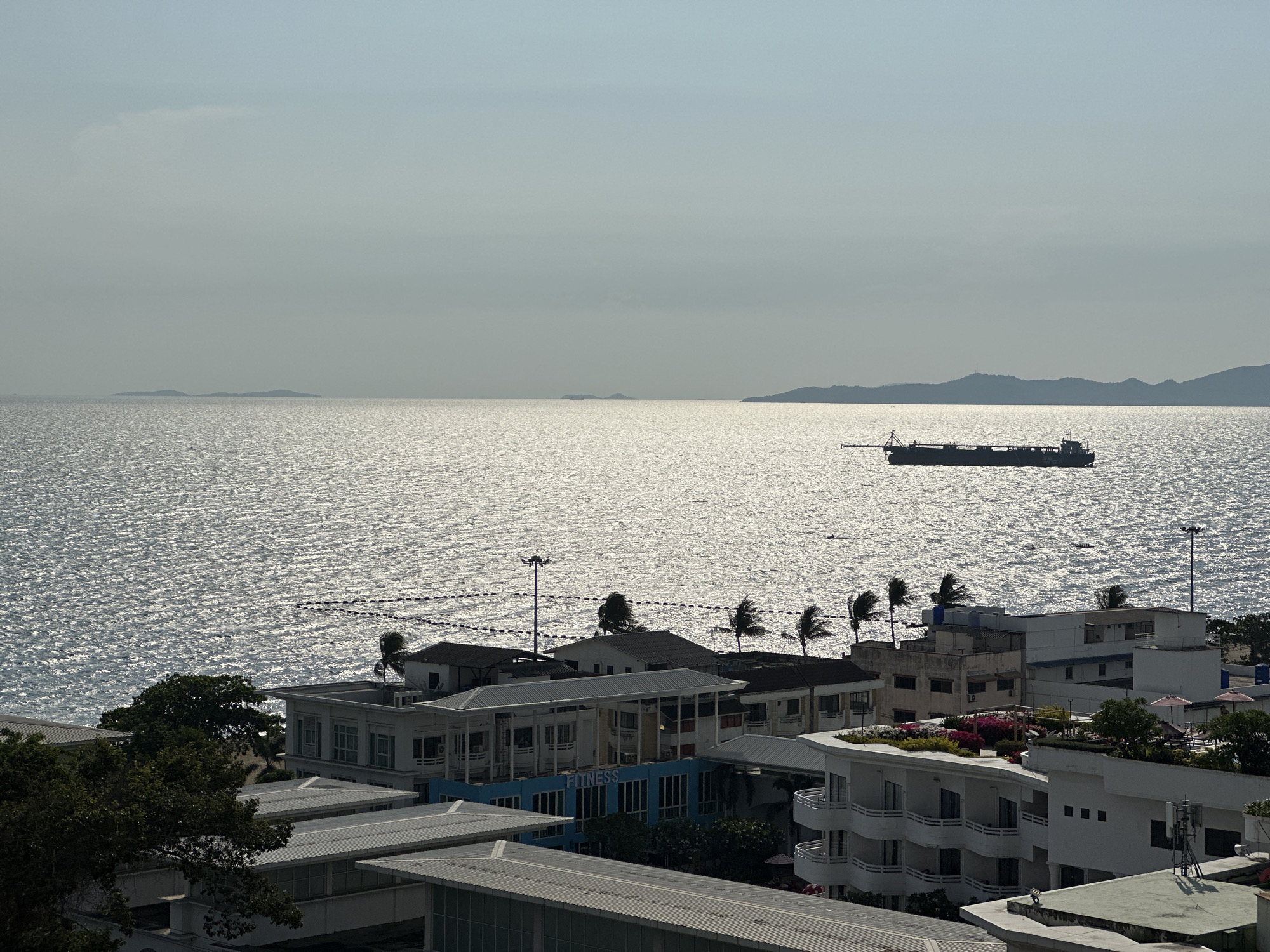 An elevated long shot captures a coastal cityscape of Pattaya City, Thailand, under a bright, hazy sky. The vast expanse of the ocean dominates the middle ground, shimmering intensely with sunlight reflecting off its surface. In the mid-distance on the water, a large, dark-hulled ship, possibly a dredge or cargo vessel, is visible, appearing somewhat static. Further out, a series of dark, low-lying islands or landforms punctuate the horizon, fading into the atmospheric haze.
In the foreground and midground, a densely packed urban landscape unfolds, featuring numerous multi-story buildings with various architectural styles, including those with balconies and flat roofs. Lush green trees are interspersed among the structures, and several palm trees, with their fronds appearing to be swayed by a breeze, add a tropical touch. On one prominent blue-colored building, the word "FITNESS" is clearly visible. Some buildings feature rooftop gardens or landscaped areas with colorful flowers. The time of day appears to be either morning or late afternoon, characterized by the low-angle sun creating the strong reflections on the water and the overall bright yet somewhat diffused lighting. The weather is bright but hazy or partly cloudy. There is no explicit human activity visible, but the bustling urban environment and the ship on the water suggest ongoing life and commerce in this vibrant coastal city.