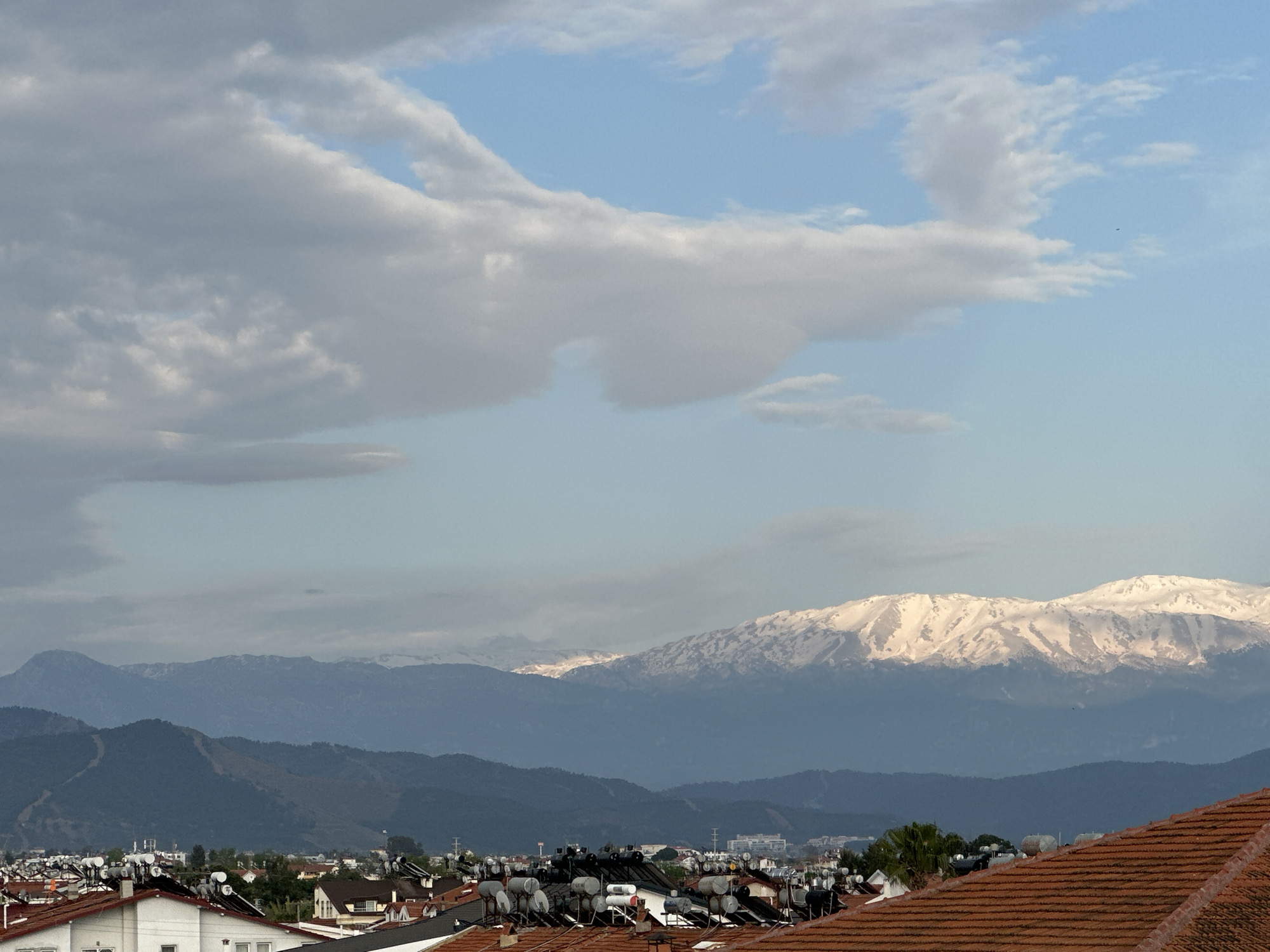 The image shows a view in Fethiye, Turkey, featuring a cityscape foregrounded by terracotta rooftops equipped with solar water heating panels. In the midground, there's a series of forested hills that transition into the snow-capped Taurus Mountains. The sky is partially covered by cumulus clouds with patches of blue visible.