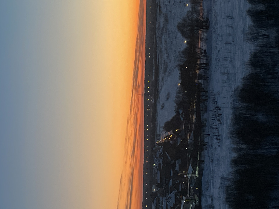 The image displays a panoramic winter landscape during twilight, likely sunset or sunrise, creating a striking contrast between the sky and the ground. The photograph appears to have been taken from a slightly elevated position, looking out over a wide expanse.
The upper portion of the image is dominated by the sky, which exhibits a beautiful gradient of colors. On the left, the sky is a soft, muted blue-grey, transitioning smoothly towards the right into vibrant oranges and yellows, indicative of the low sun near the horizon. There are subtle horizontal streaks or wisps of darker clouds within the orange part of the sky, adding texture.
A distinct horizontal line marks the distant horizon, separating the colorful sky from the darker terrestrial features. Just below this horizon line, a thin band of land is visible, dotted with numerous small, bright yellow-orange lights, suggesting a distant road or a very spread-out settlement.
The lower half of the image depicts a snow-covered ground, bathed in the cool light of twilight. The snow appears a pale blue-grey, reflecting the ambient light. On the left side of the midground, a cluster of buildings, possibly houses or a small village, is visible. Their roofs are dark, likely covered in snow, and several warm, artificial lights glow from within or around these structures, casting small pools of illumination. Further into the midground and extending towards the right, the landscape opens into what appears to be a vast, snow-covered field or plain. Scattered across this snowy expanse are numerous dark, bare trees and bushes, their silhouettes stark against the lighter ground. Some trees stand individually, while others form denser clusters, particularly along the right edge of the frame. The overall impression is one of cold serenity, with the warm hues of the sky providing a dramatic backdrop to the muted, wintry scene below.
There is no discernible activity or visible text in the image.