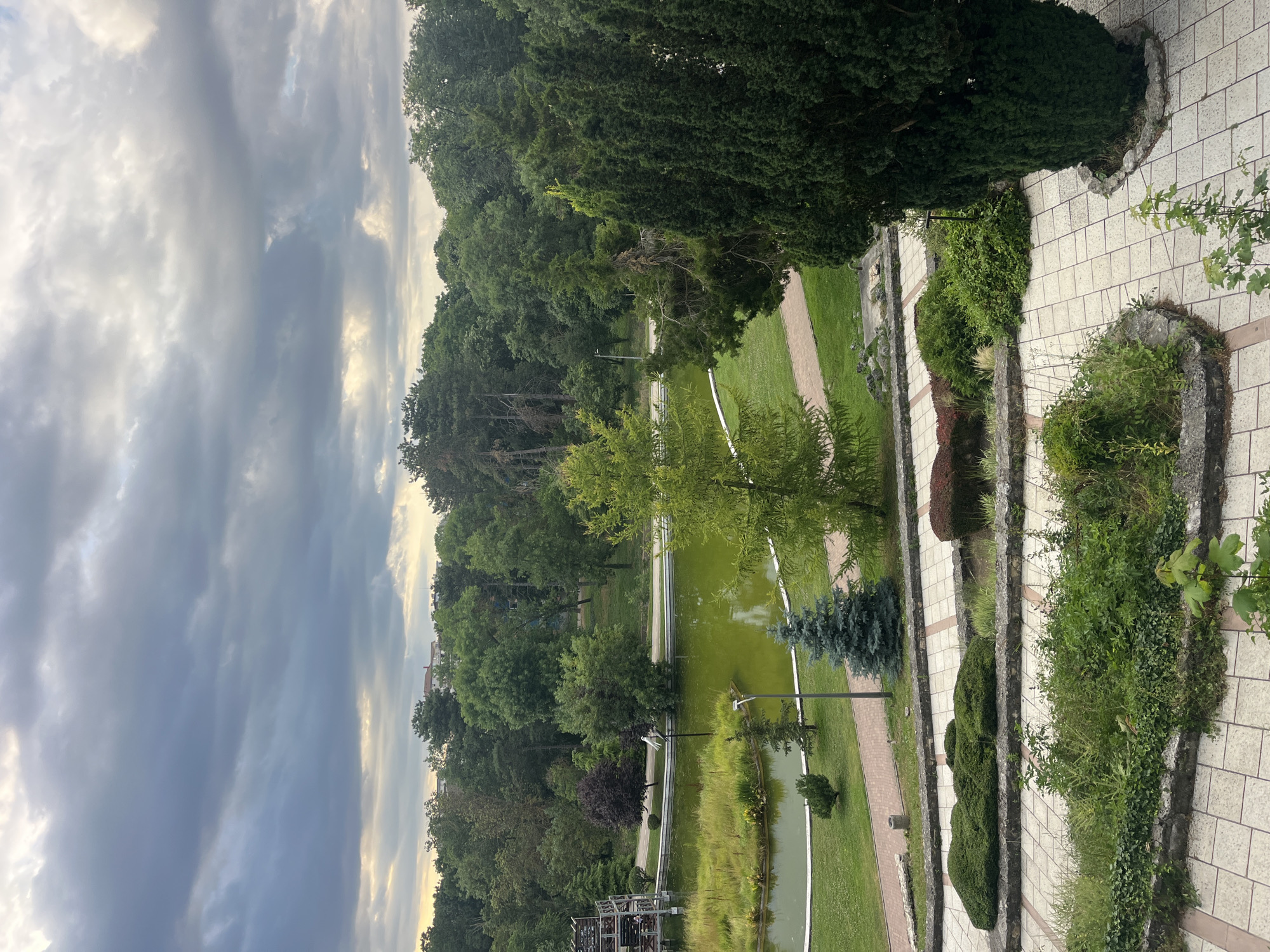 The image shows a park in Dobrich, Bulgaria. In the foreground, there are paved walkways and low, manicured bushes. Beyond this, a body of green water, likely a pond or lake, is visible, with grassy banks and a path running alongside it. Several trees are scattered around the water, including a tall, dark green conical tree on the right and a lighter green deciduous tree in the middle. In the distance, a dense line of trees forms a backdrop to the park. A modern, multi-story structure, possibly a building or part of a leisure facility, is partially visible on the far left, adjacent to the water. The sky above is filled with dramatic, cloudy formations. There are no people visible in the frame.