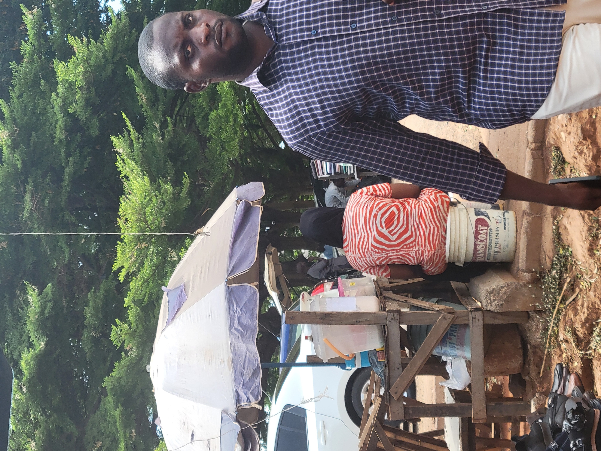 A man in a plaid shirt stands near a makeshift wooden stand topped with an umbrella, possibly a street vendor in Abuja, Nigeria. The stand holds containers and a bucket labeled "TRANSCOAT." A person in an orange and white patterned top sits behind the stand. A white car is parked nearby. The scene appears to be outdoors, with trees in the background and exposed earth around the stand.