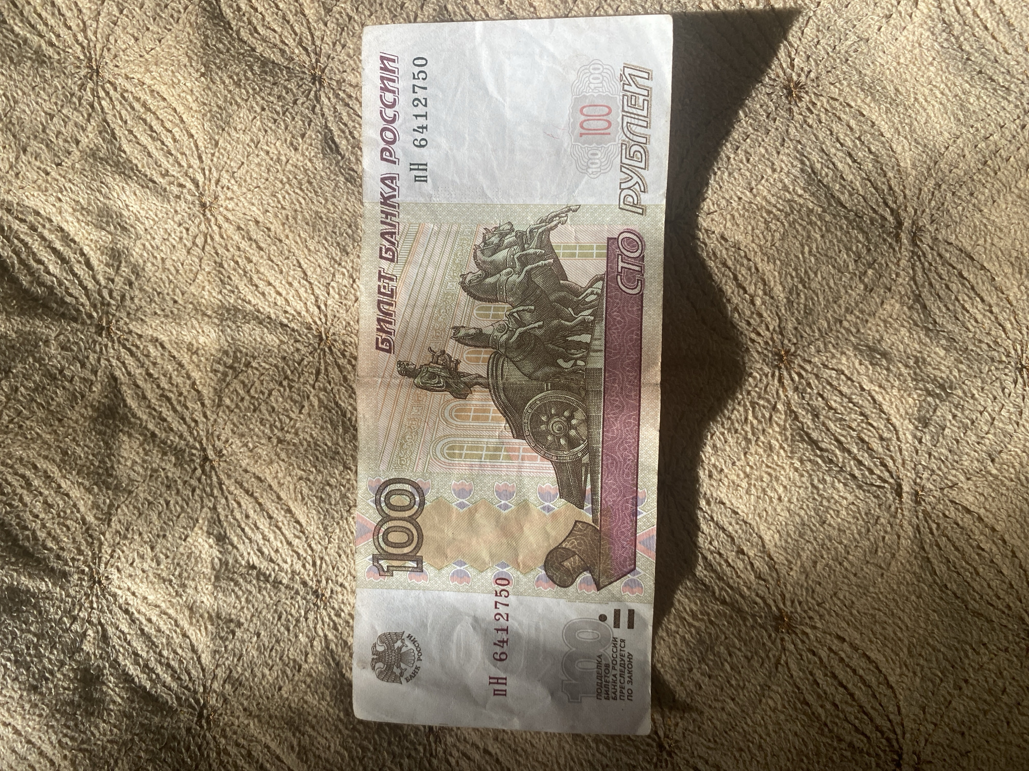 The image shows a Russian 100 ruble banknote lying on a textured brown fabric surface. The banknote is oriented horizontally and is illuminated by sunlight, casting a shadow to the bottom right. The front of the banknote is visible, featuring the number "100" prominently on the left and right sides, along with Russian text and an image of a quadriga on the facade of the Bolshoi Theatre. The serial number "pH 6412750" is also visible. The fabric surface appears to be a blanket or upholstery with a raised, quilted pattern. There are no people or specific landmarks of Kizilyurt, Russia visible in this image; the image solely focuses on the banknote and its immediate background.