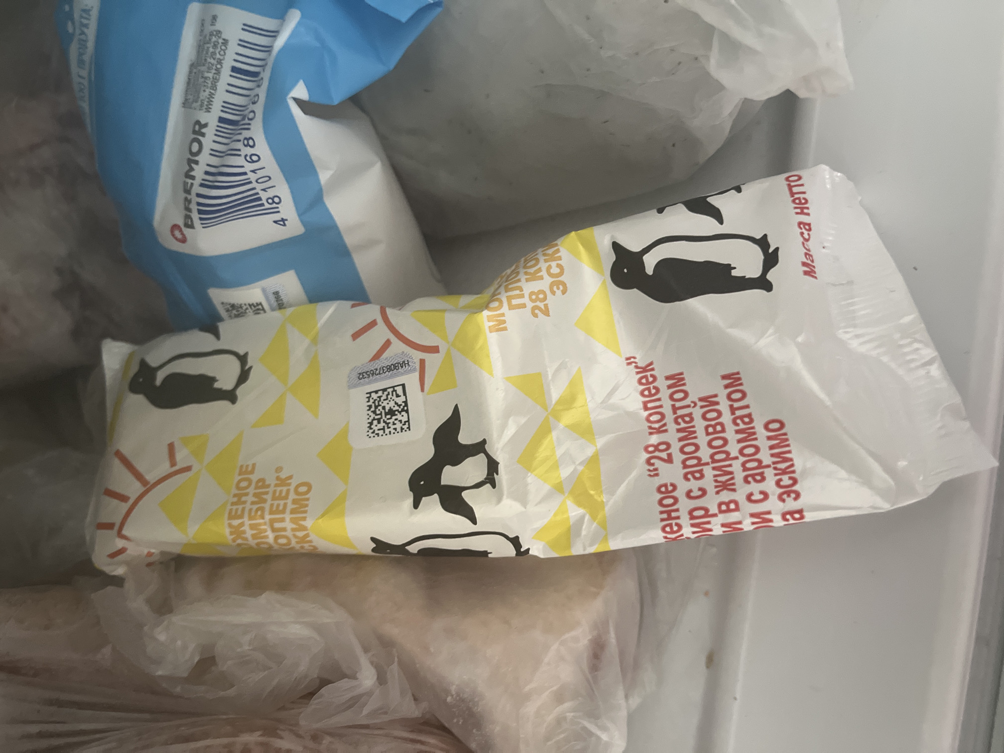 The image is a close-up, top-down shot of frozen food items in a freezer. The primary subject is a white plastic wrapper with yellow triangular patterns and black penguin illustrations. Text on the wrapper is in Russian and appears to be product information, including "ЖЕННОЕ" (likely part of "мороженое" - ice cream), "28 КОПЕЕК" (28 kopecks, a denomination), and "ЭСКИМО" (eskimo, a type of ice cream bar). There are also some QR codes and serial numbers visible on the wrapper. To the right of this wrapper is a blue plastic bag with a barcode and text in both Russian and English, including the brand name "BREMOR." In the background, other frozen items are visible, partially obscured by plastic bags. The overall environment is that of a freezer, indicated by the frost and the frozen nature of the products. The scene is indoors, in a domestic or commercial refrigeration unit. There are no people or animals present, nor any indication of a specific location within Minsk, Belarus beyond the possibility that the products are manufactured or sold there.