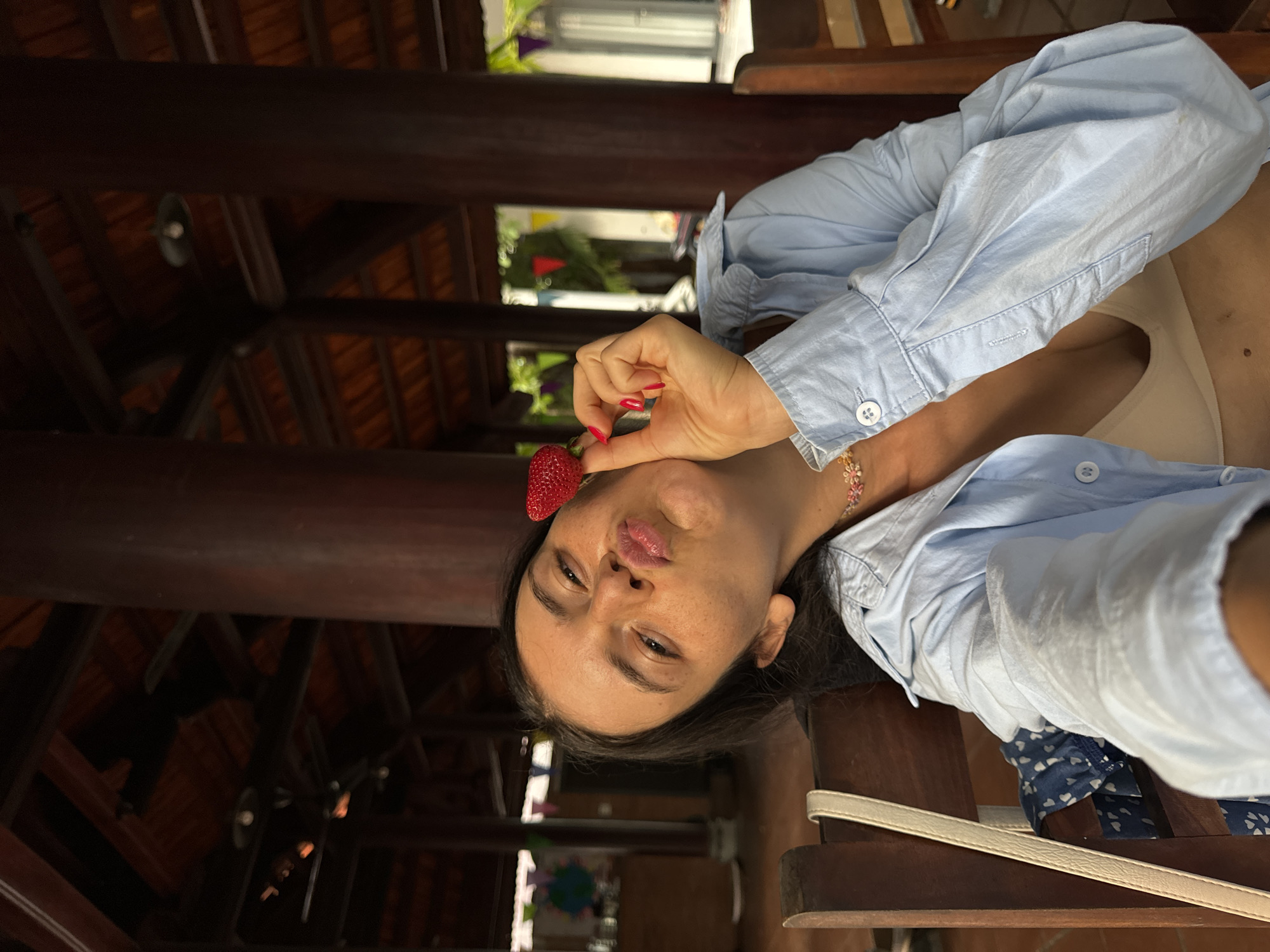 The image shows a young woman posing for a selfie in a tropical setting, likely Mũi Né, Vietnam. She is holding a strawberry to her lips, making a playful pout. The woman is wearing a light blue button-down shirt over a cream-colored tank top and has on a delicate necklace. Her nails are painted a vibrant pink.
The background reveals a wooden structure, suggesting an outdoor patio or a covered seating area with a thatched roof. Beams and rafters are visible overhead, characteristic of traditional Southeast Asian architecture. Sunlight filters through, indicating it is daytime, likely warm and bright given the tropical location. In the distance, glimpses of lush greenery and possibly water can be seen, reinforcing the outdoor, possibly beach-adjacent, nature of the scene. The overall mood is relaxed and joyful. No text is visible in the image.