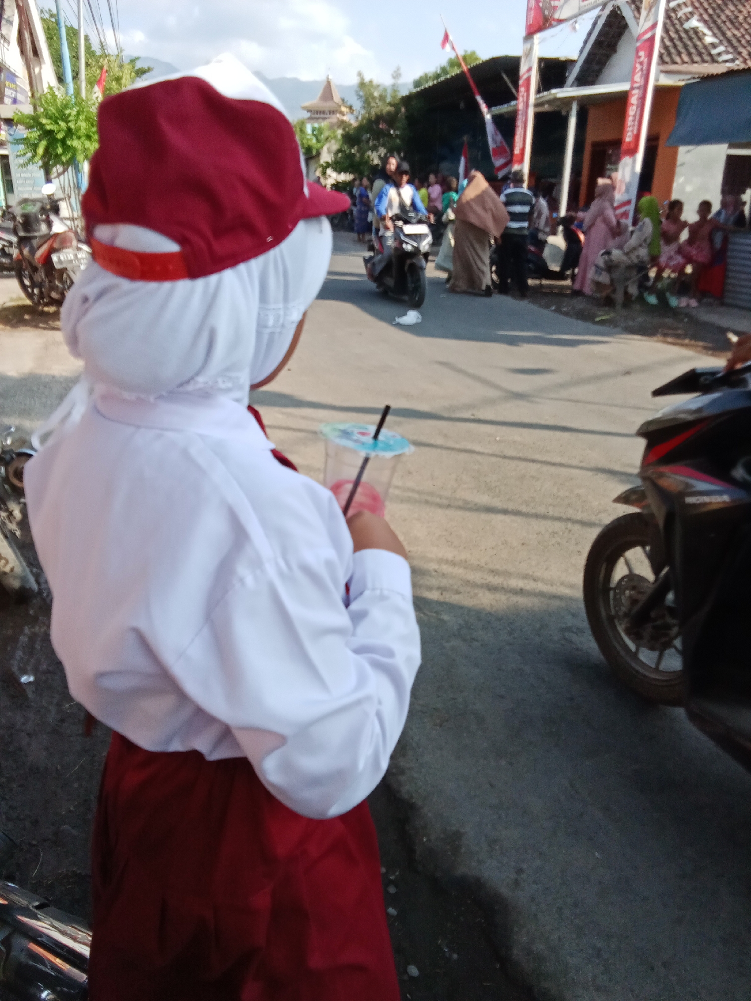 In Bungatan, Indonesia, a young person wearing a school uniform, a white hijab, a white shirt, and a maroon skirt is standing in the foreground, holding a plastic cup with a straw. They are facing away from the camera, looking towards a street lined with buildings and people. The street appears to be busy with pedestrians and motorcycles. In the background, there are decorations, including banners and flags, suggesting a local event or celebration. The environment is an outdoor urban setting with a paved road and buildings on either side. The sky is visible, with some clouds.