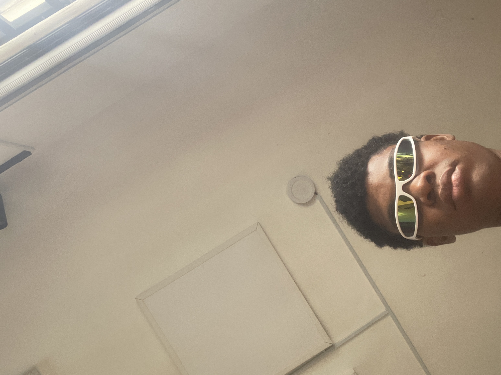 A low-angle, close-up shot shows the upper half of a person wearing white, wraparound sunglasses with reflective lenses. The person has dark, curly hair and dark skin. The background is a plain, light-colored wall, possibly a ceiling, with a rectangular panel on the upper left and a circular light fixture in the upper center. The lighting suggests an indoor environment, possibly during the day with natural light coming from the right. No specific location in Ado, Nigeria can be identified from the image.