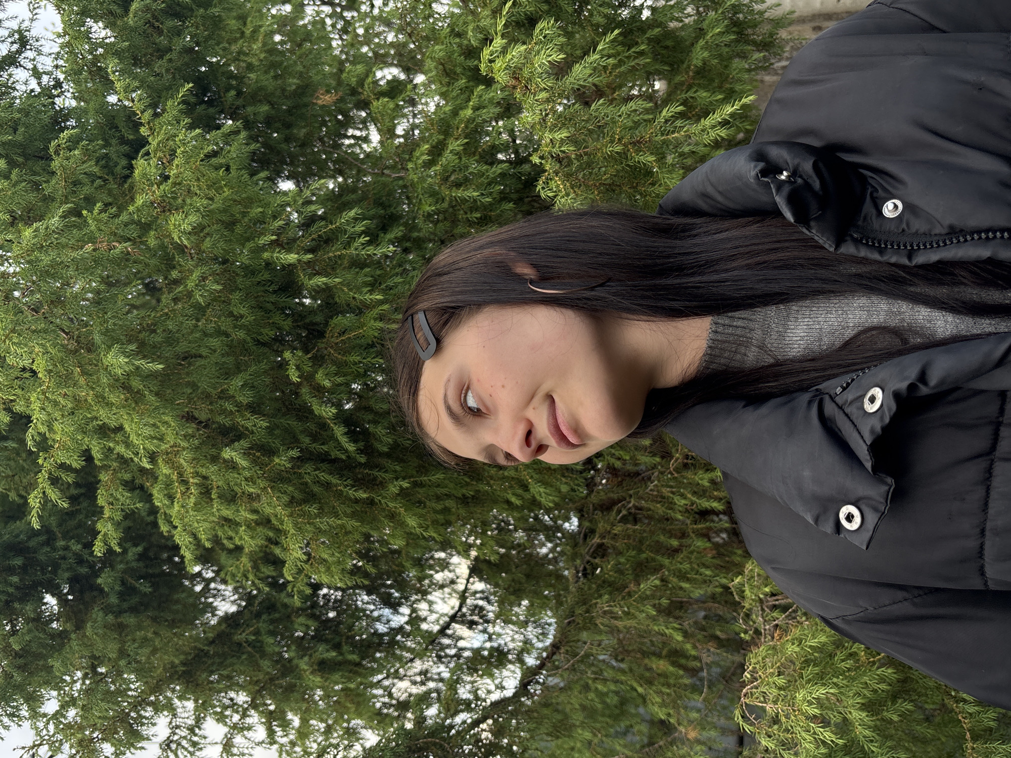 A young female, appearing to be in her late teens or early twenties, is depicted outdoors in a waist-up shot. She has long, dark brown hair, with a black rectangular hair clip positioned on the right side of her head. A thin, dark-colored hoop earring is visible in her right ear. Her complexion is light, with visible freckles on her upper cheek and nose. She wears a grey crew-neck knit sweater beneath a black puffer jacket featuring a high collar and metallic snap fasteners on the front and collar. Her gaze is directed upwards and to the left, with a subtle smile on her lips. The background is dominated by dense, vibrant green coniferous foliage, likely a juniper or similar evergreen tree. Patches of light, overcast sky are visible through the gaps in the branches. The scene is set in the city of Фоки (Foki), Russia.