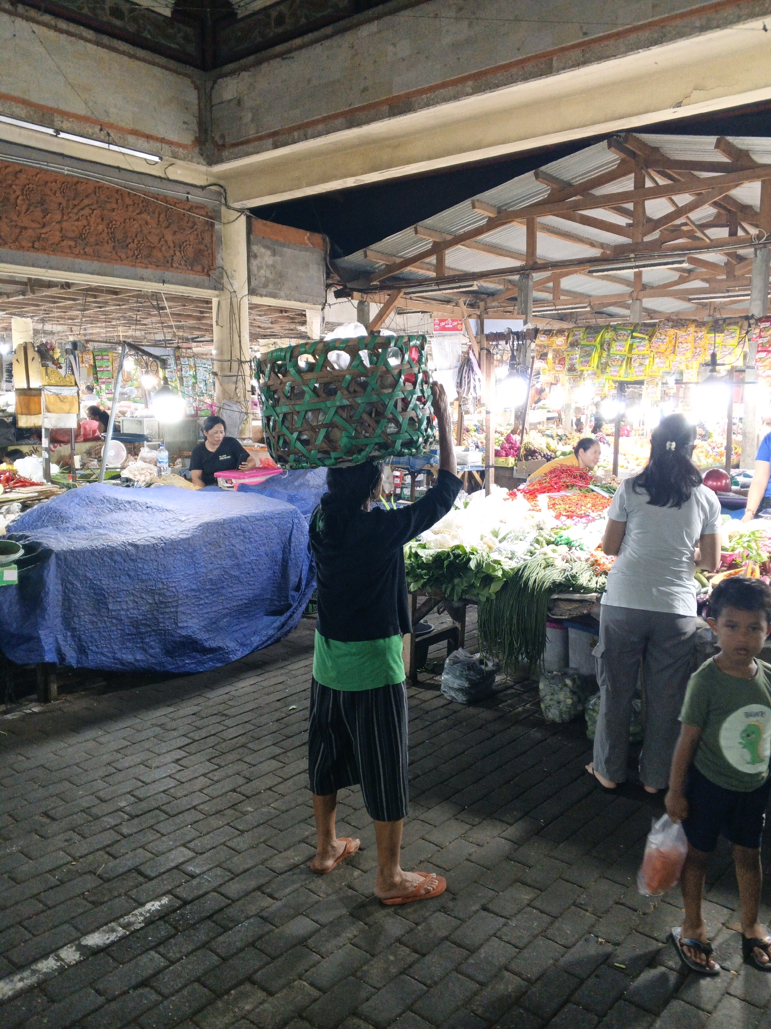This is an exterior, eye-level, full-shot photograph of a market stall in Denpasar, Indonesia. The scene depicts a busy marketplace with an array of fresh produce and goods. In the foreground, a person wearing a black shirt, green vest, and striped pants walks away from the camera, carrying a large woven basket on their head. To the right, a young boy in a green t-shirt and dark shorts walks forward, holding a plastic bag with orange contents. A woman in a white shirt and grey pants stands behind a stall laden with vegetables, facing away from the camera. Another woman in a black t-shirt is seated behind a table covered with a blue tarp, to the left of the frame. The market appears to be covered with a corrugated metal roof supported by wooden beams, and illuminated by fluorescent lights. The ground is paved with dark interlocking bricks. The background shows more stalls with various food items and packaged goods. The overall atmosphere suggests a typical night market scene in Denpasar.