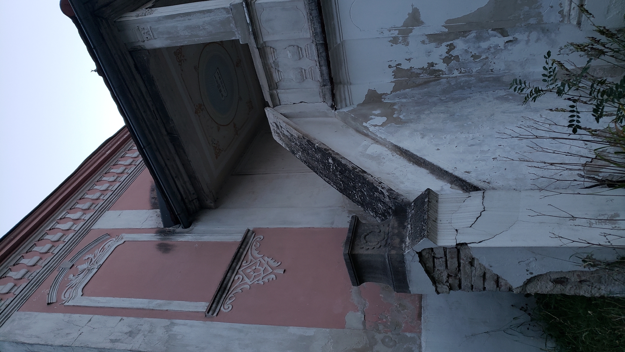 The image shows a close-up of the exterior of an old, dilapidated building, likely a house, with a pink facade and white ornate details. A weathered staircase, also showing signs of decay, leads up to a covered porch with decorative elements on its ceiling. The building's walls are cracked and peeling, and bricks are exposed in some areas, indicating neglect. Overgrown vegetation, including dry branches and some green leaves, is visible around the base of the structure. The sky is a pale blue, suggesting it might be early morning or late evening. The scene conveys a sense of abandonment and decay. This building is located on Južná trieda in Prešov, Slovakia.