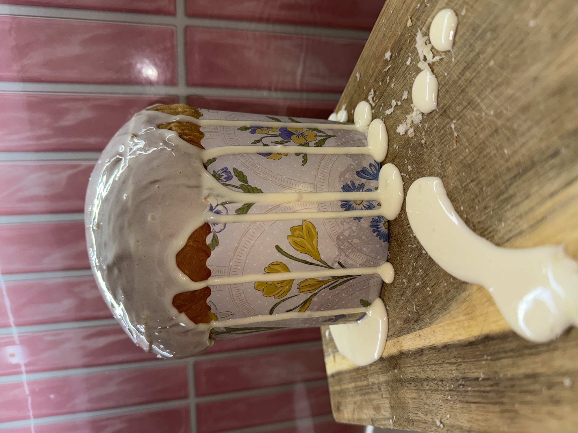 The image shows a baked pastry, possibly a kulich, covered in white icing. The pastry is decorated with a floral paper wrapper and sits on a wooden board. White icing drips down the sides of the pastry and is also pooled on the board. The background features a pink-tiled wall. The image was taken in Krasnoyarsk, Russia.