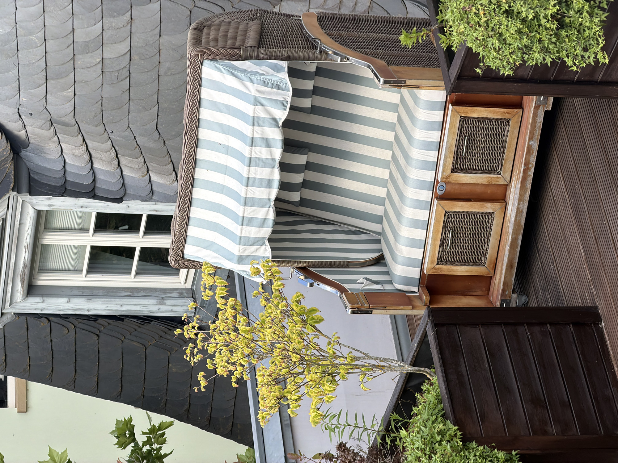 The image depicts an outdoor scene on a wooden deck, likely a balcony or terrace, in Meisenheim, Germany. The focal point is a woven beach chair with blue and white striped upholstery. The chair has two small, woven-front storage compartments at its base and sits on wheels. Beside the chair, a small tree with yellowing leaves is planted in a dark brown wooden planter box, which is filled with dark soil and some greenery. Another dark brown wooden planter box with lush green foliage is visible on the right side of the chair. In the background, a building features a dark slate-tiled roof and a window with white mullions and blinds. The slate tiles are arranged in an overlapping, scalloped pattern, characteristic of some traditional German architecture. The environment appears to be calm and residential. There are no people or animals visible in the image.