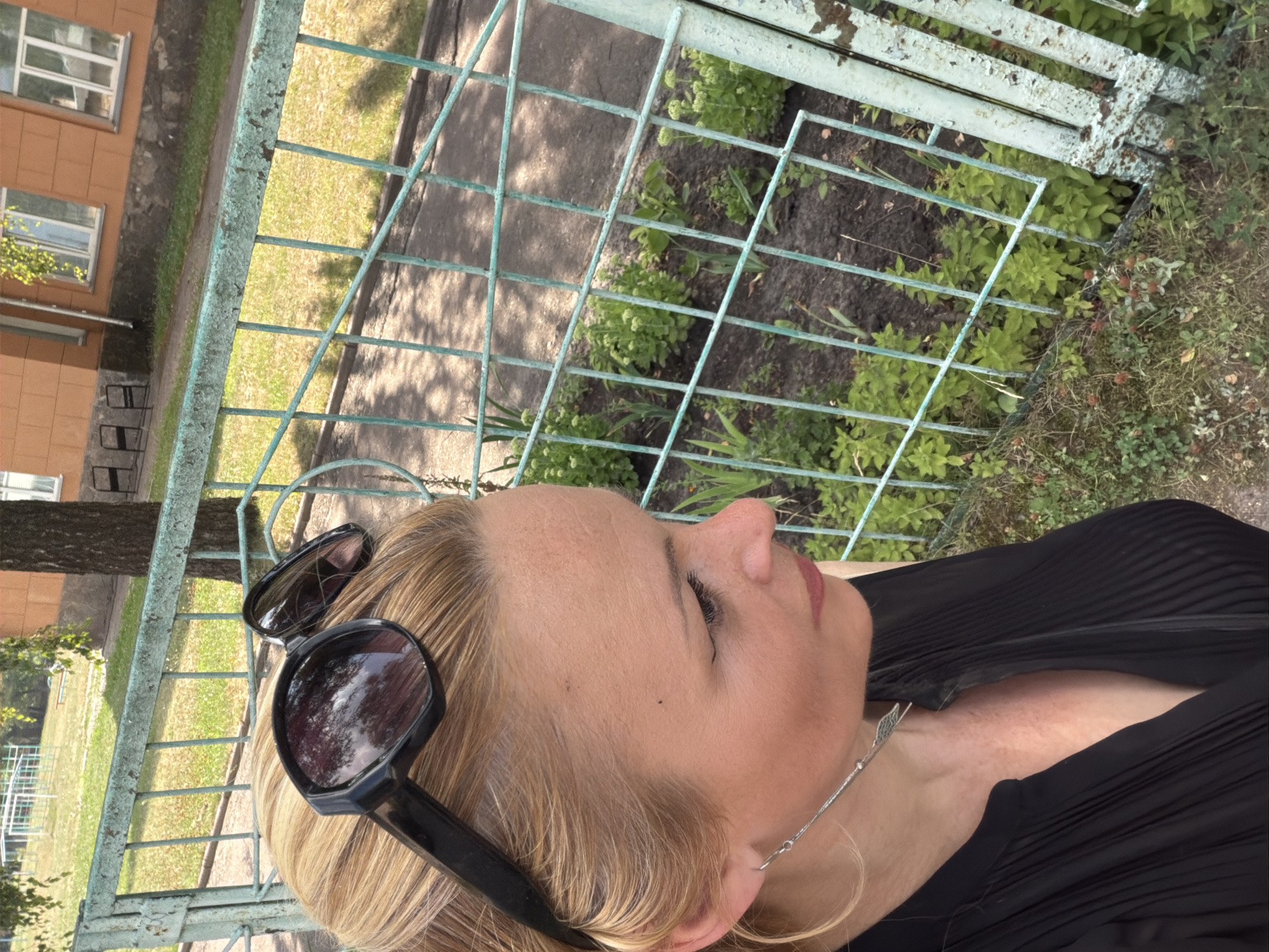In a medium-angle, outdoor, close-up shot, a woman with blonde hair, wearing a black sleeveless top and sunglasses, stands in front of a distressed, pale green metal fence. Her head is slightly tilted, and she is looking to the right. Behind her, the fence encloses a patch of greenery. The background includes a building with an orange facade, a grassy area with a playground structure, and trees. The lighting suggests it is daytime.