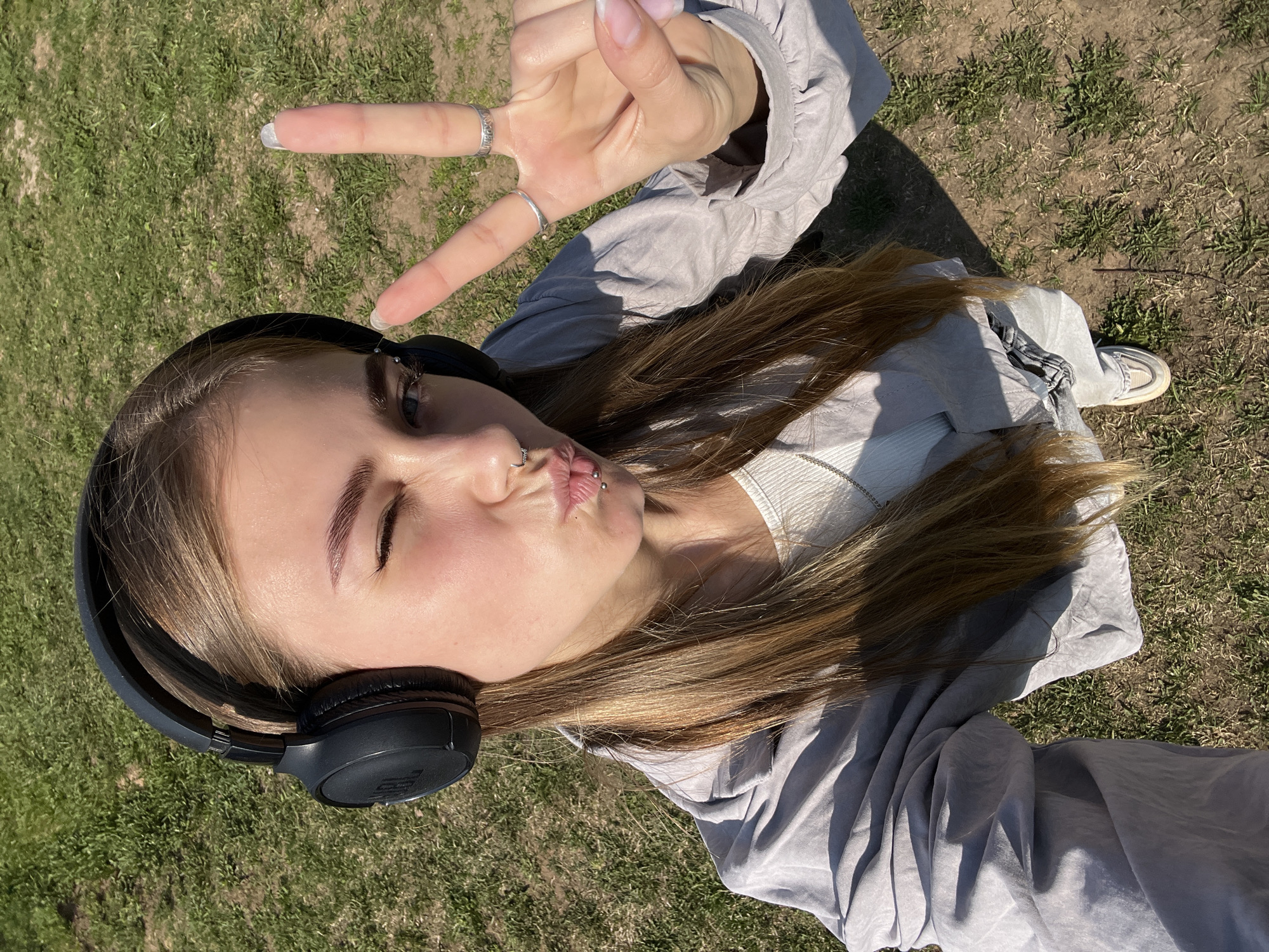 A selfie of a female with long light brown hair, headphones, piercings and making the peace sign, is set in a grassy outdoor location. The woman, wearing a gray shirt over a white top, tilts her head and winks while puckering her lips. She wears black headphones and has multiple piercings on her nose and lips. Her hand shows several silver rings. She wears white shoes. The background consists of grass, suggesting a park or open space. The image is taken from a high angle. The presence of green grass and sunlight indicates a daytime setting.