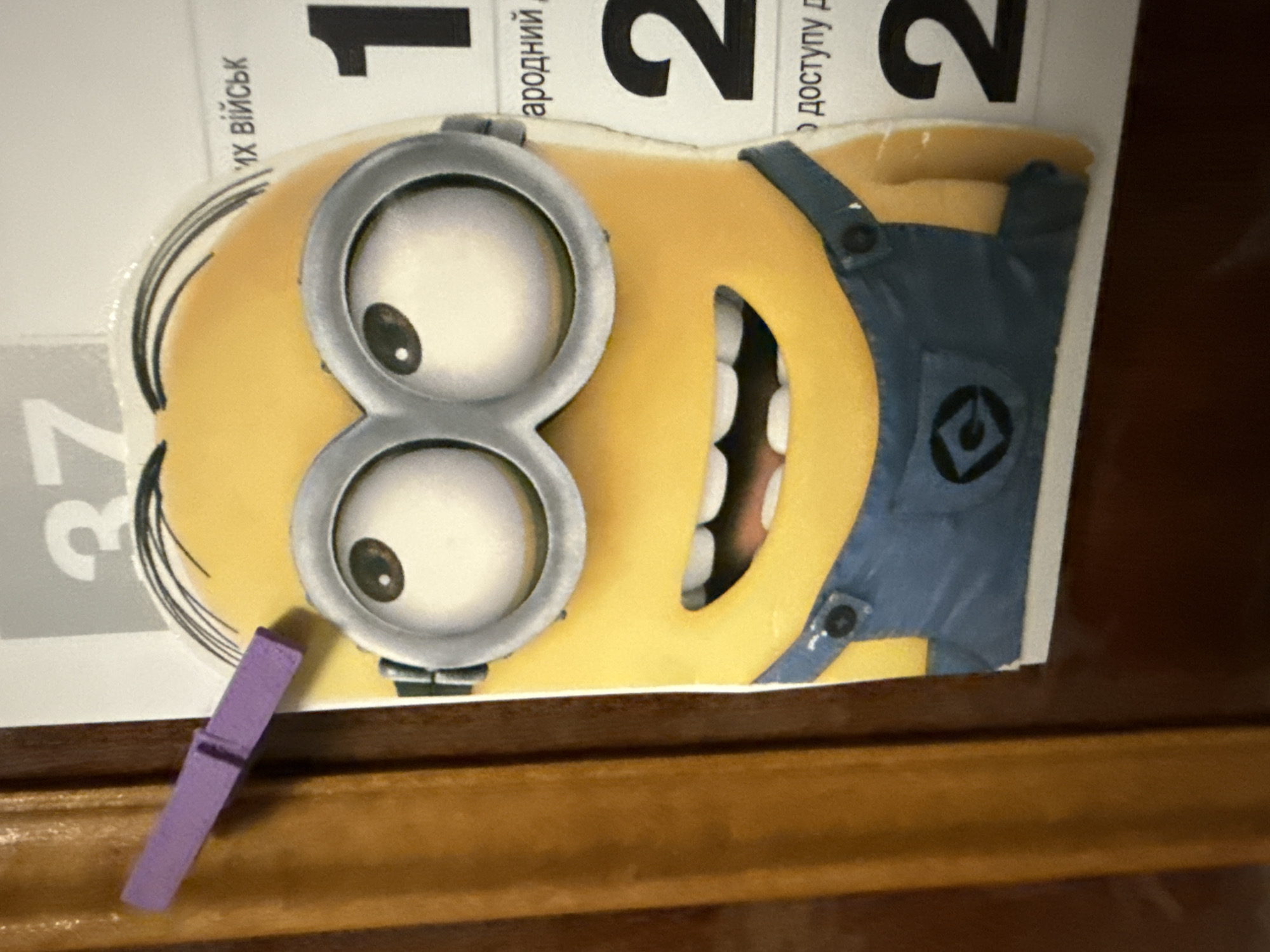 The image shows a close-up of a Minion character sticker, attached to a surface by a purple clothespin on the left. The Minion is yellow with large goggles and a blue overalls. Behind the Minion, there is a portion of a calendar or poster with Ukrainian text and large black numbers "1" and "2". The background to the left appears to be a wooden surface, possibly a door or furniture. The lighting is bright, casting a slight shadow on the Minion's face. The location is unknown.