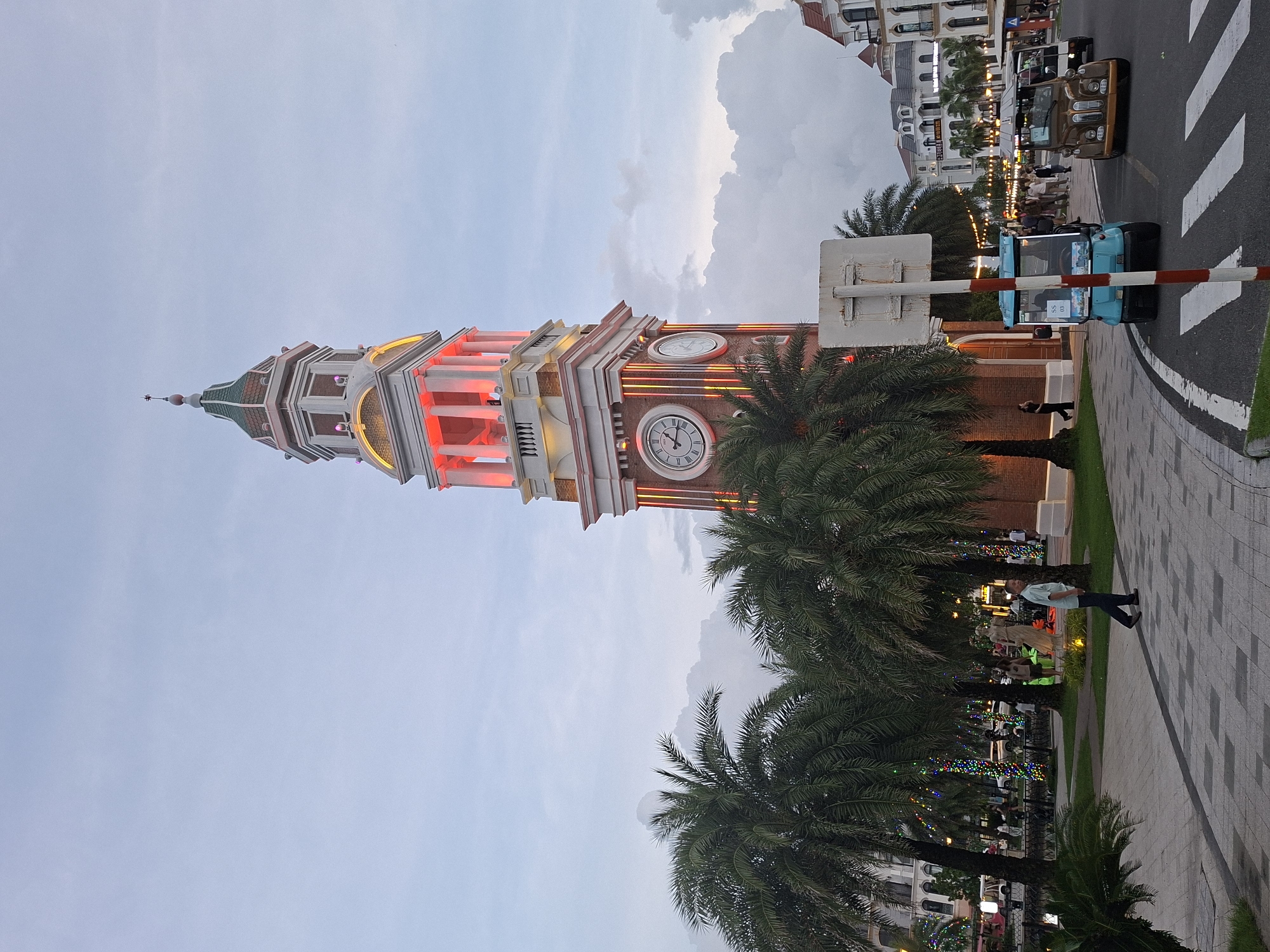 This media file is an outdoor shot of a clock tower and its surroundings, likely in Phú Quốc, Vietnam. The clock tower is illuminated with red and yellow lights, suggesting it is either dusk or nighttime. The sky is overcast with large clouds, and the overall lighting is soft.
The scene features the tall, multi-story clock tower with visible clock faces. Surrounding the tower are palm trees, some with colorful string lights draped around them, indicating a festive atmosphere. A paved walkway with a diamond pattern runs alongside the scene, and a street with a pedestrian crossing is visible in the background.
Several people are present in the scene. One person stands on the walkway, looking up at the tower, appearing to be a tourist. Another person walks on the sidewalk, and there are more individuals in the background near buildings and streetlights. Two vehicles, a brown jeep-like vehicle and a blue golf cart-style vehicle, are on the street.
The presence of palm trees, the style of the buildings in the background, and the warm weather implied by the outdoor setting with people in light clothing all align with a location in Vietnam like Phú Quốc. The lighting and decorations suggest a leisure or entertainment area, perhaps a resort or a town square. The overall mood is relaxed and pleasant, with the illuminated tower serving as a focal point. There is a sign on the blue vehicle that reads "SS 03".