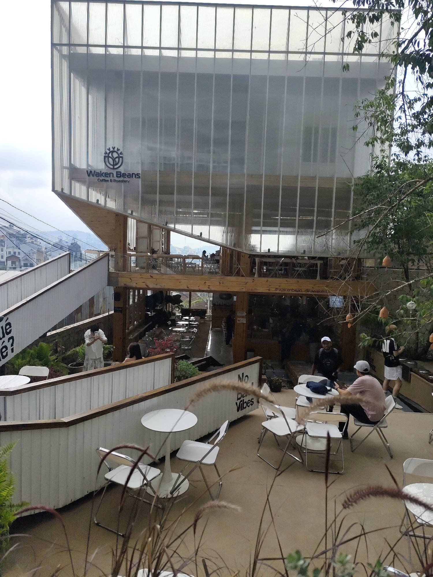 The image shows an outdoor seating area of "Waken Beans Coffee & Roastery" in Da Lat, Vietnam. The multi-level structure of the café is visible, with a translucent top section. Tables and chairs are arranged on different levels, with patrons seated or standing nearby. A partially obscured staircase leads to higher levels. Various plants and vegetation are present in the foreground and around the building. The sky is overcast.