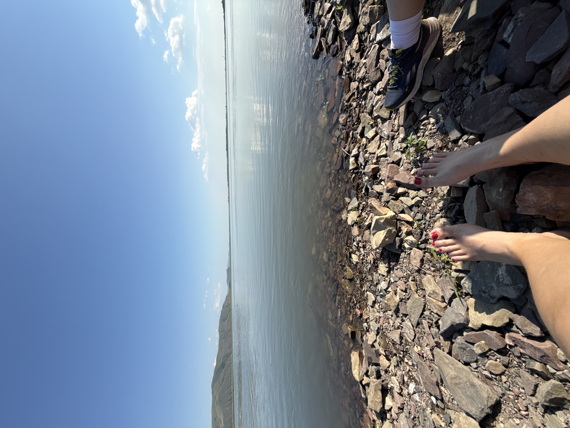 In the city of Ust'-Abakan, Russia, two individuals are seated on a rocky shore beside a wide, calm body of water under a clear blue sky with scattered white clouds. The foreground is dominated by numerous stones and rocks of varying sizes and colors, indicating a natural, unpaved shoreline.
One individual, on the left, is positioned with their legs extended towards the water. Their bare feet are visible, with toenails painted red. The skin tone suggests warm weather.
The second individual, on the right, is partially visible, wearing a dark athletic shoe with yellow laces and white socks. Their legs are also resting on the rocks.
The water stretches out towards a distant horizon, where low, undulating hills form a muted backdrop. The surface of the water is smooth with gentle ripples and reflects the sky. A thin, dark twig or branch lies partially submerged in the shallow water near the shore. The overall scene suggests a peaceful outdoor setting, possibly a lake or riverbank, during daylight hours.