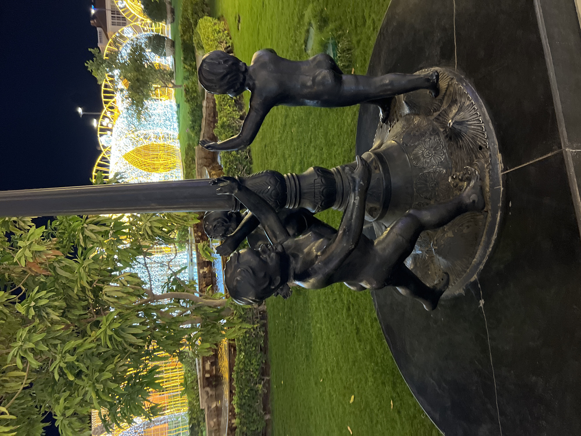 The image shows a bronze statue of several cherubs gathered around a lamppost base at night. The statues are dark and detailed, depicting children in various playful poses. The base of the lamppost is ornate, with carved patterns. The ground around the statue is covered in neatly manicured green grass. In the background, there are large, illuminated structures made of white and yellow lights, creating an archway effect. A lush green tree with many leaves is visible to the left of the statues. The lighting suggests it is an outdoor public space, likely decorated for an event or holiday. The scene is set in Sharm El Sheikh, Egypt, and appears to be a nighttime view of a well-lit recreational or entertainment area.